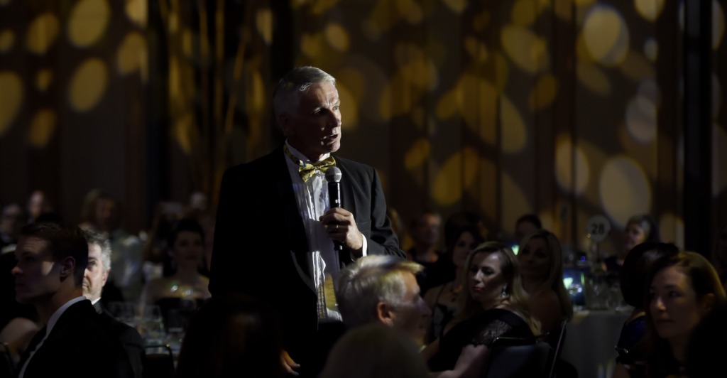 Man in a suit with a yellow bow tie speaks into a microphone at a formal event.