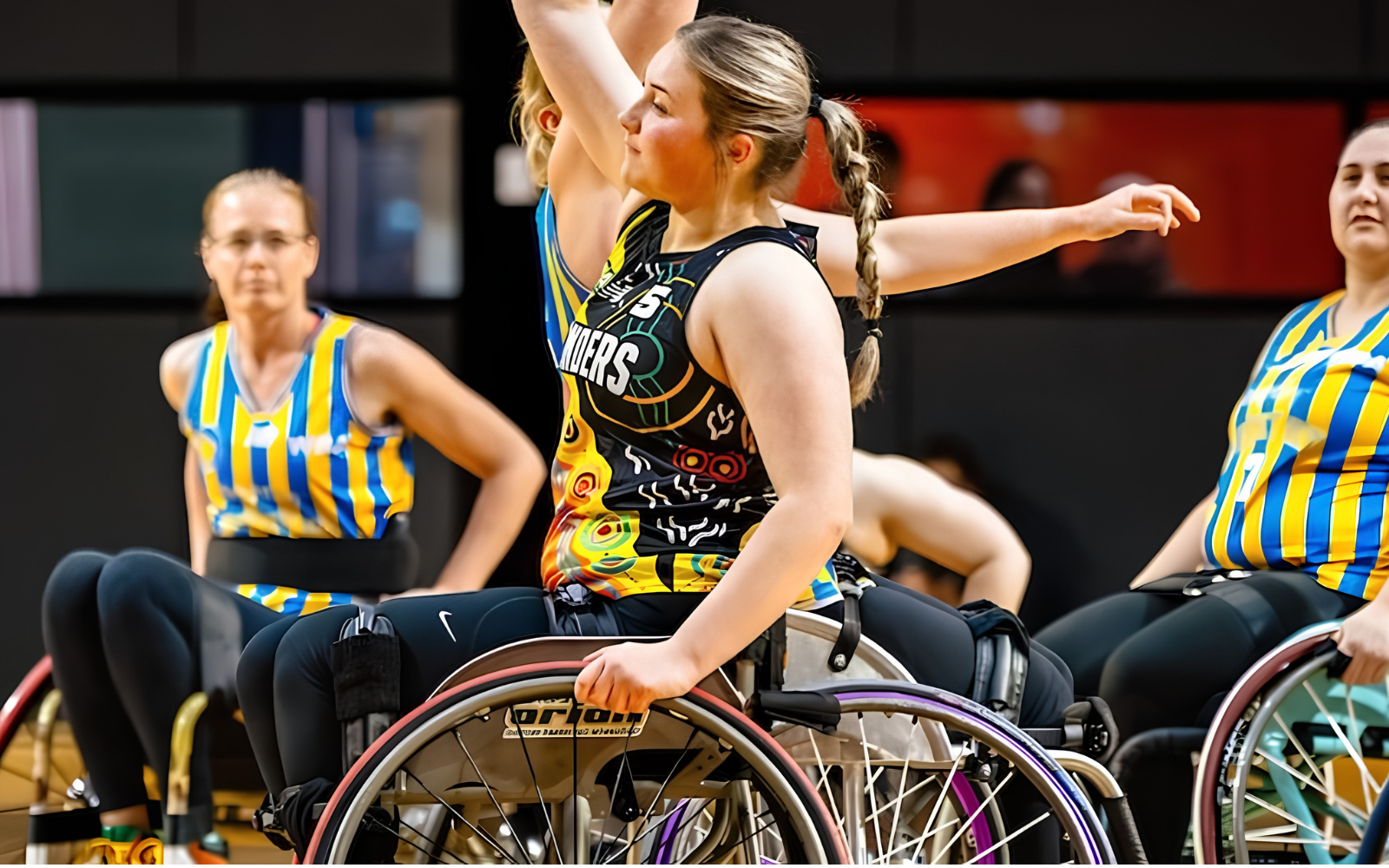 Female wheelchair rugby player in a colorful jersey raises her arm during a match, with teammates in striped uniforms nearby.