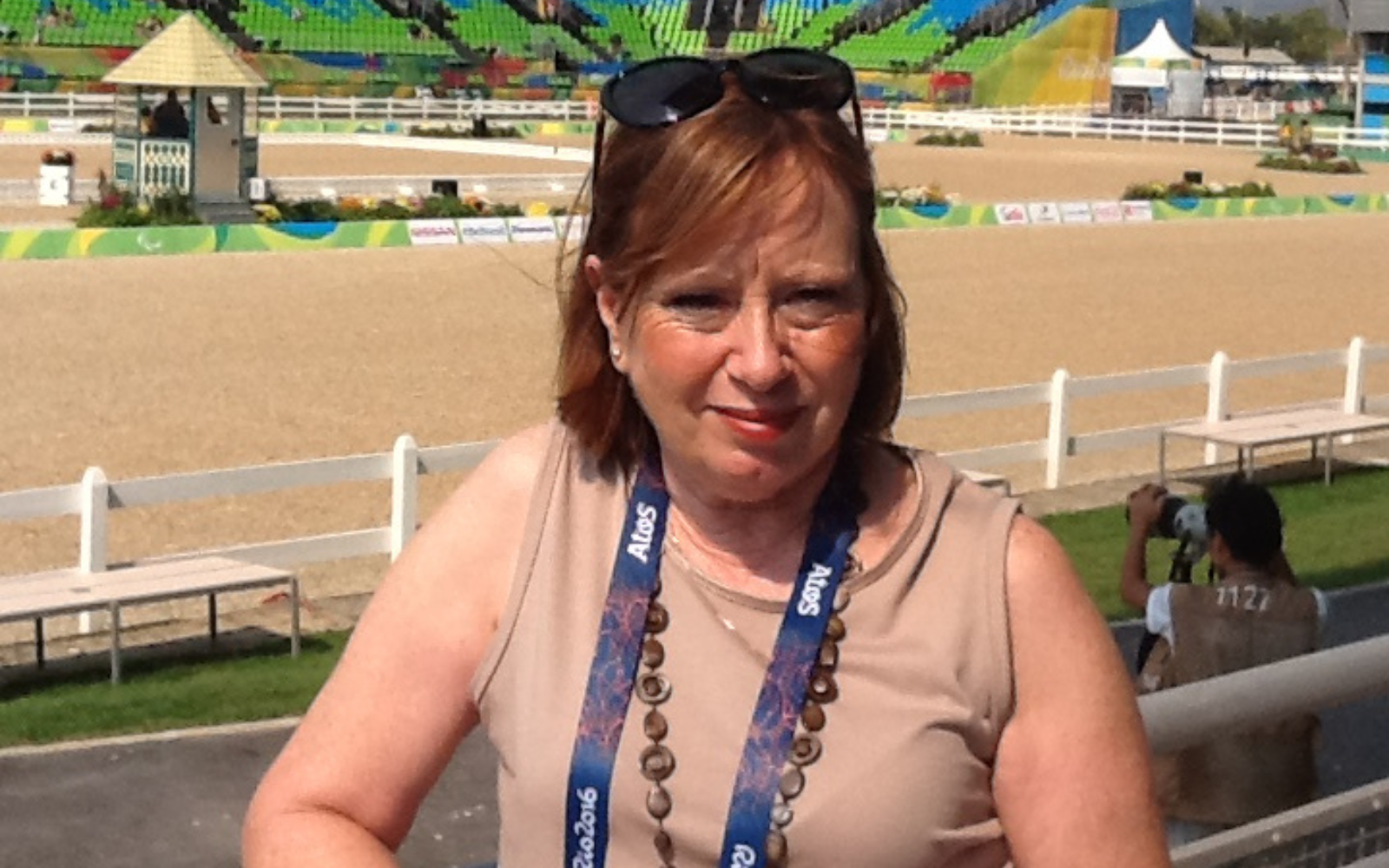 Middle-aged woman with short light brown hair and sunglasses on her head, wearing a beige sleeveless top and beaded necklace, at an outdoor arena background.