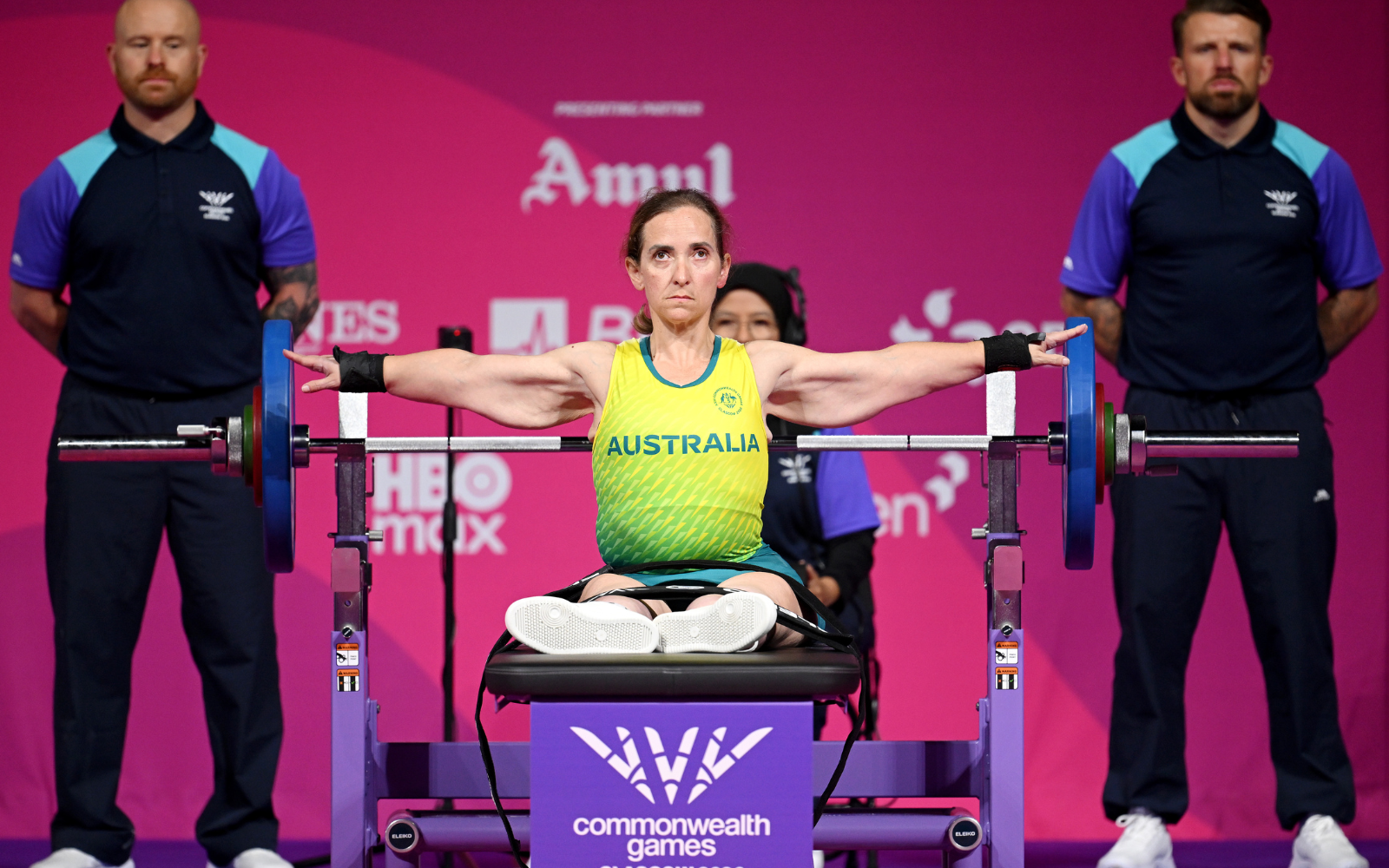 Australian para powerlifter seated on a bench, arms extended holding a loaded barbell during a Commonwealth Games event.
