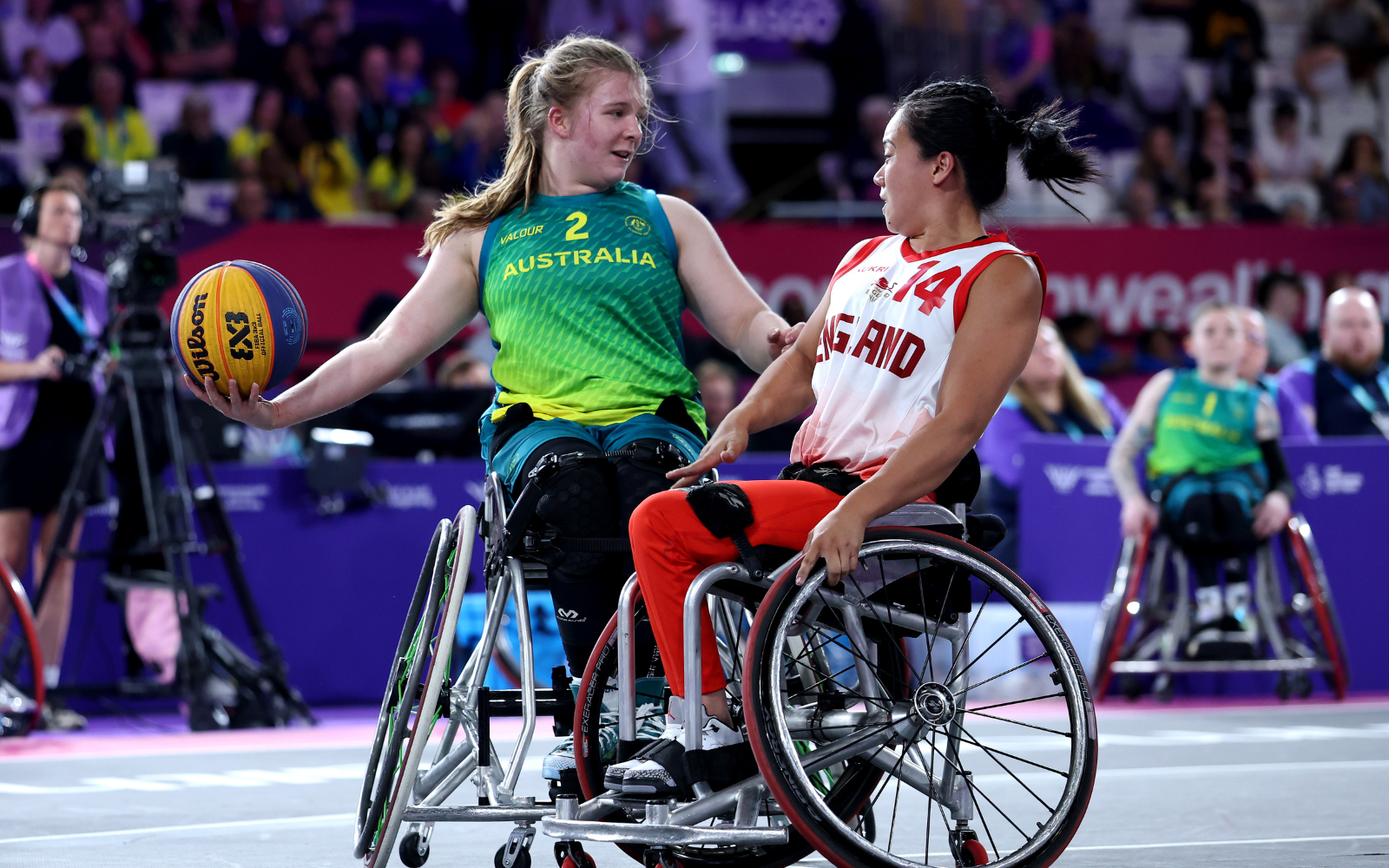 Australian player in green-yellow jersey (number 2) passes a ball to an England opponent in a wheelchair rugby match with a crowd in the background.