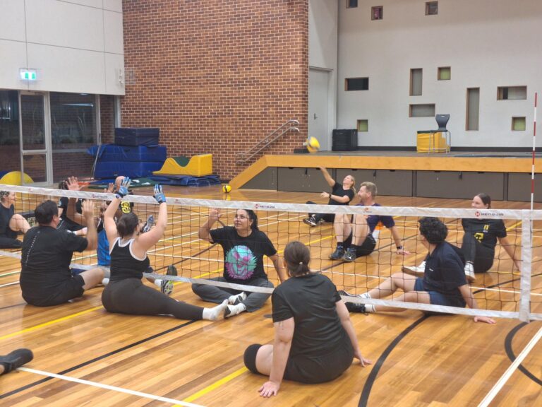 Indoor gym volleyball game with players seated on the wooden court near the net, reaching up as a yellow volleyball is hit in the air.