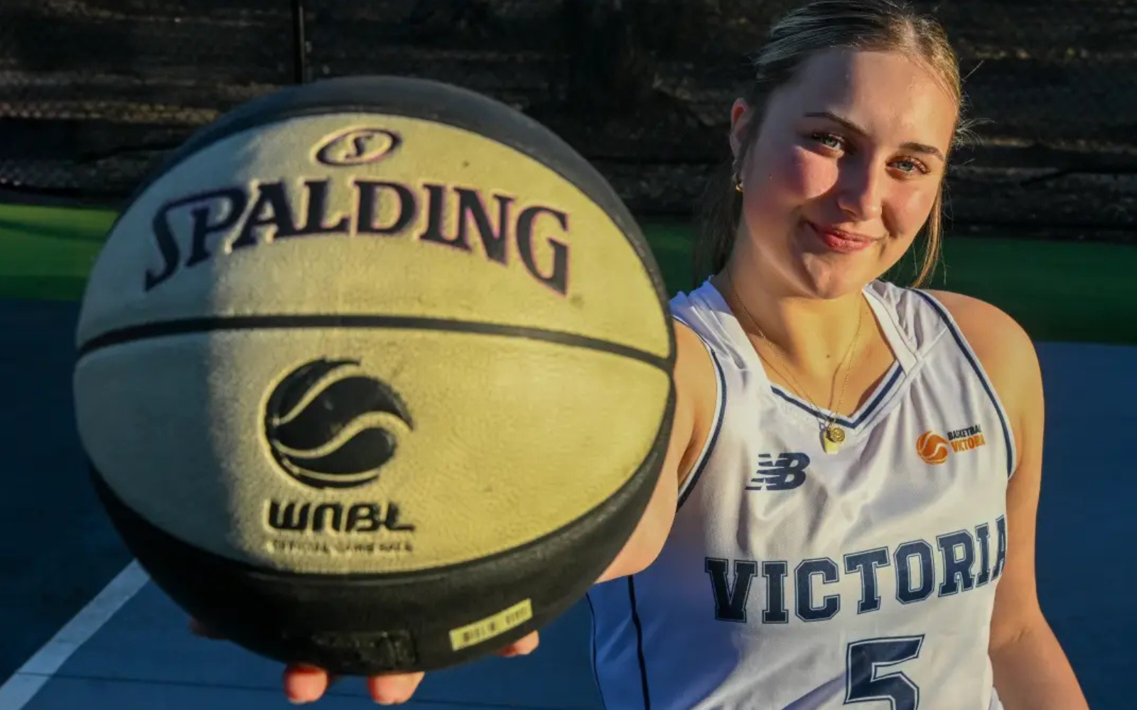 Female basketball player in a white Victoria jersey extends her arm toward the camera, holding a Spalding basketball.