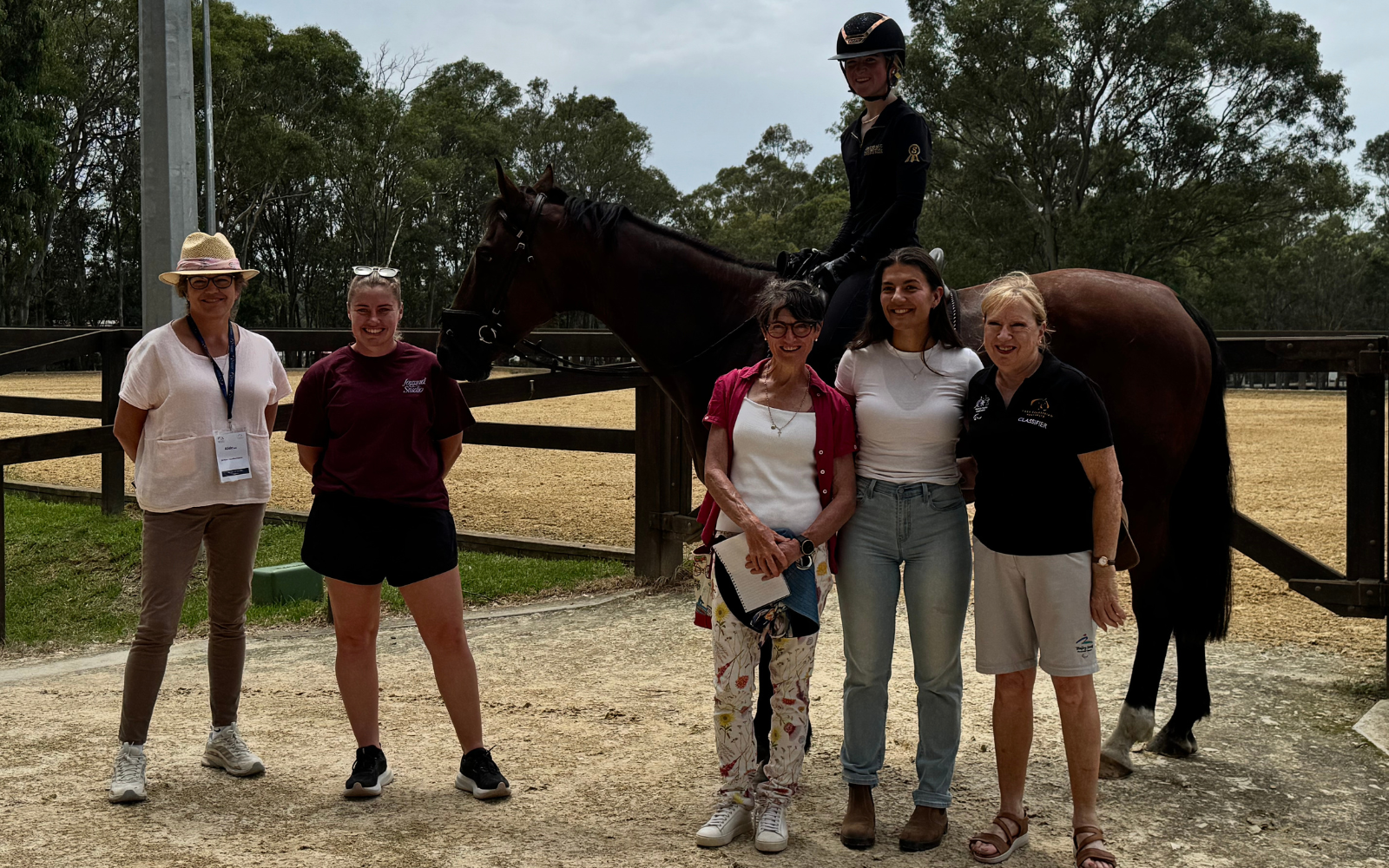 Six adults posing in an outdoor paddock; a rider sits on a dark horse behind them near a wooden fence.