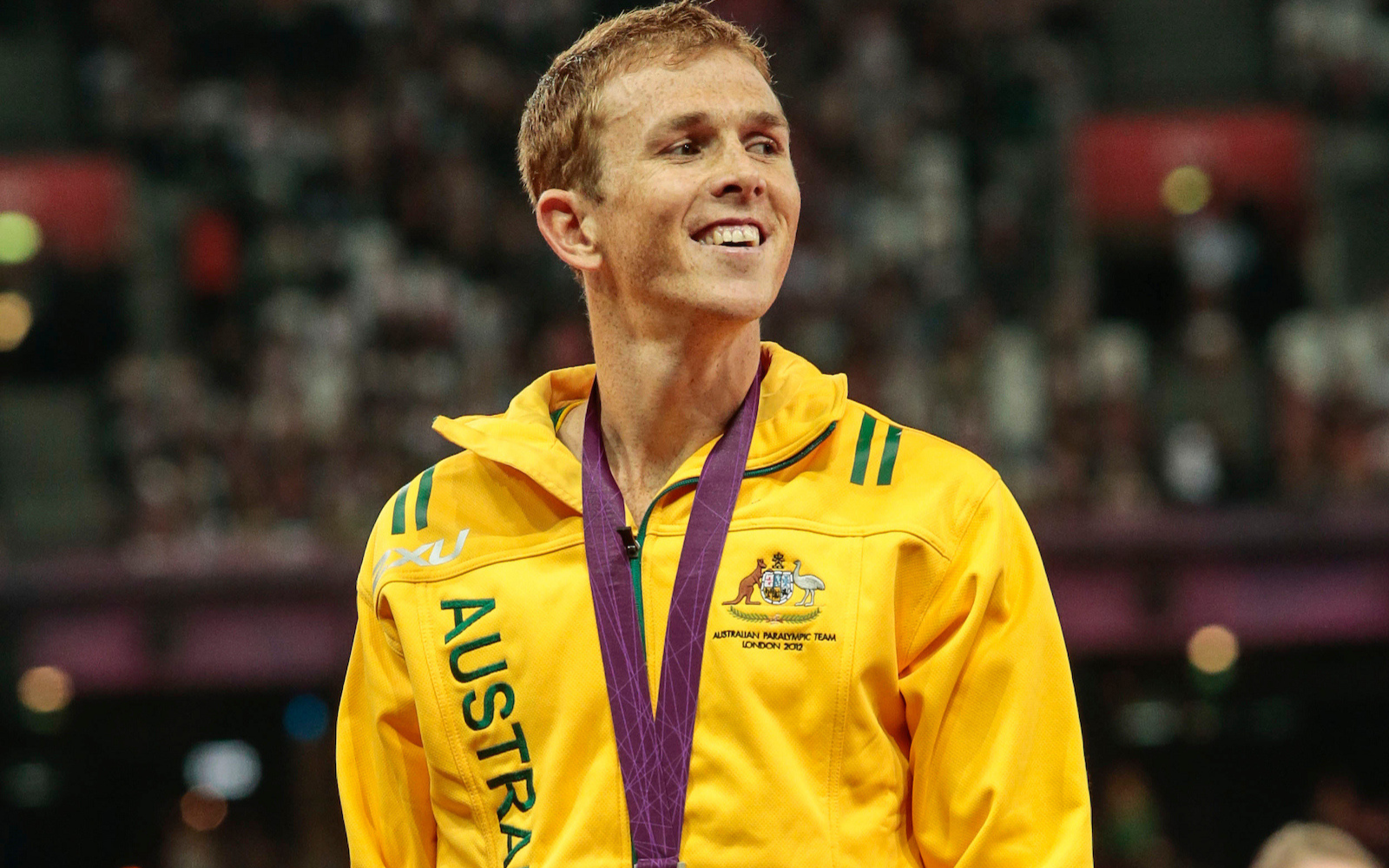 Australian Paralympic athlete in a yellow jacket with a purple medal ribbon after the event, smiling on stage.