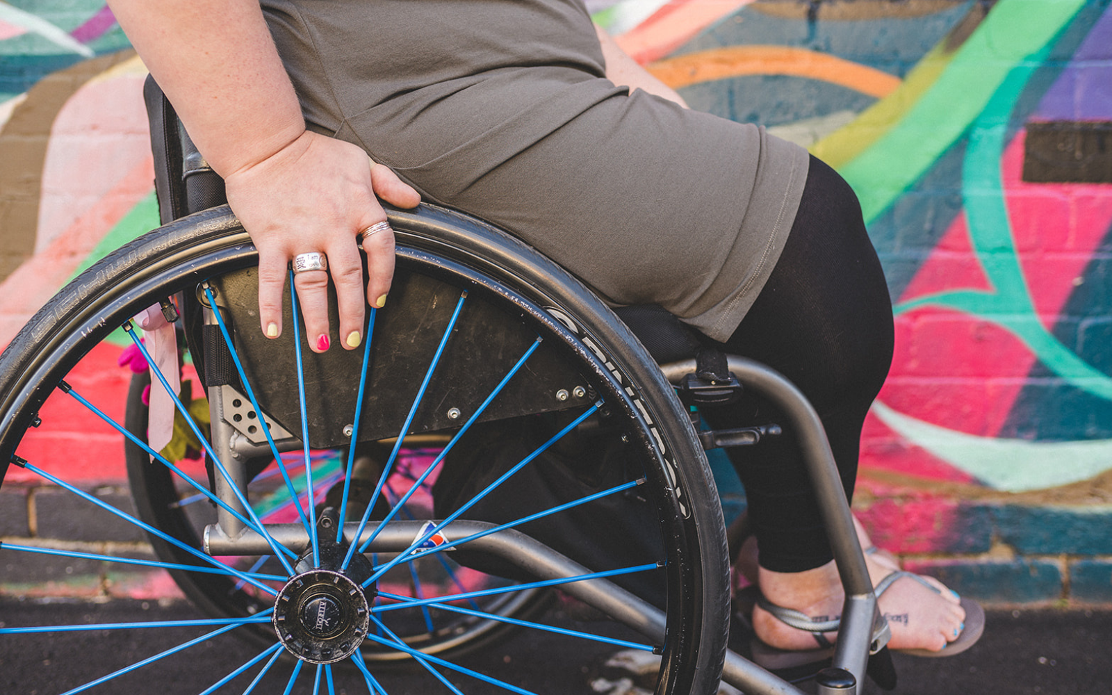 Person in a wheelchair resting their left hand on the blue-spoked wheel, with a colorful graffiti wall behind them.