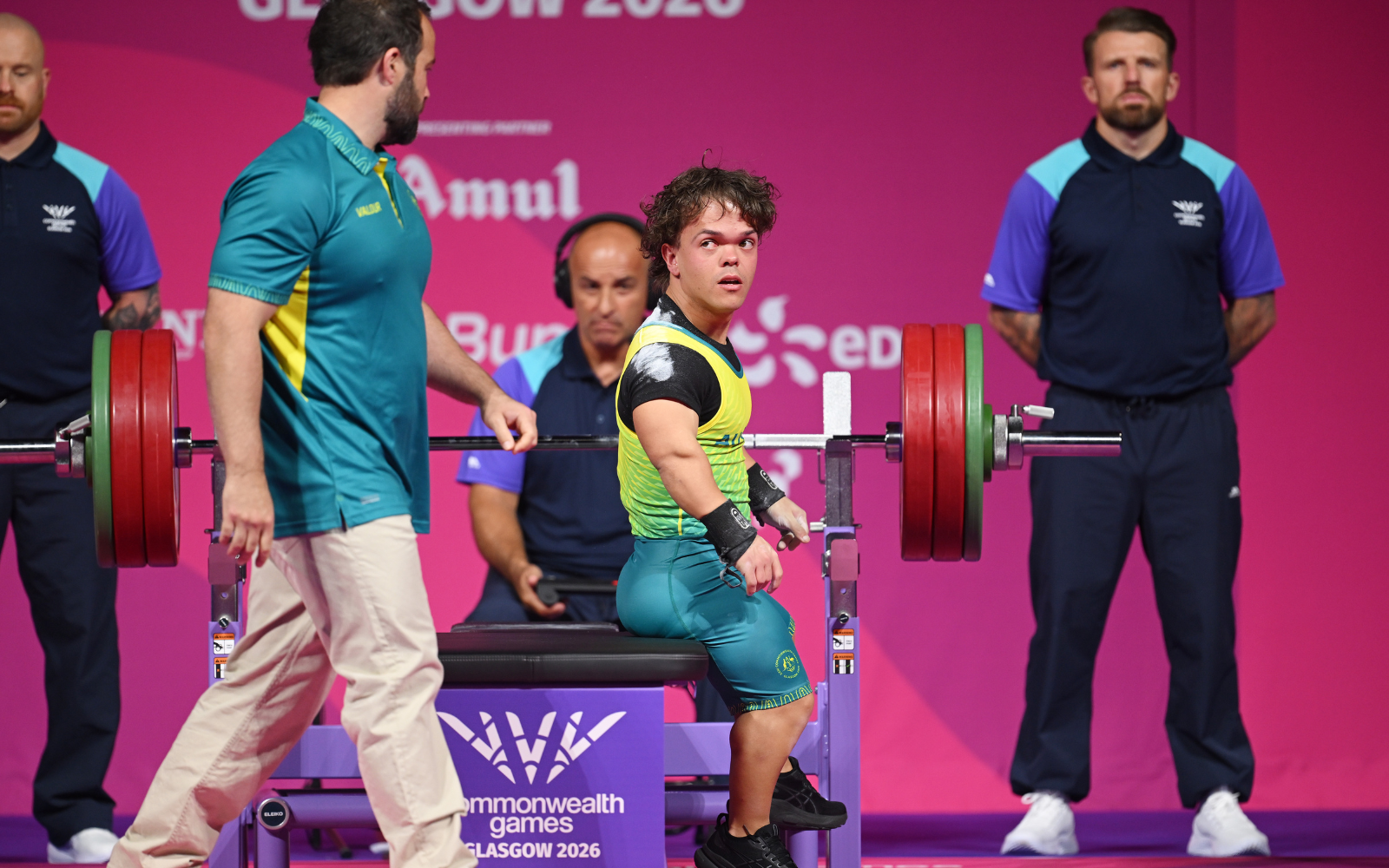 Male weightlifter in a neon-yellow top and teal shorts sits on a bench with a loaded barbell, flanked by two spotters against a pink backdrop.