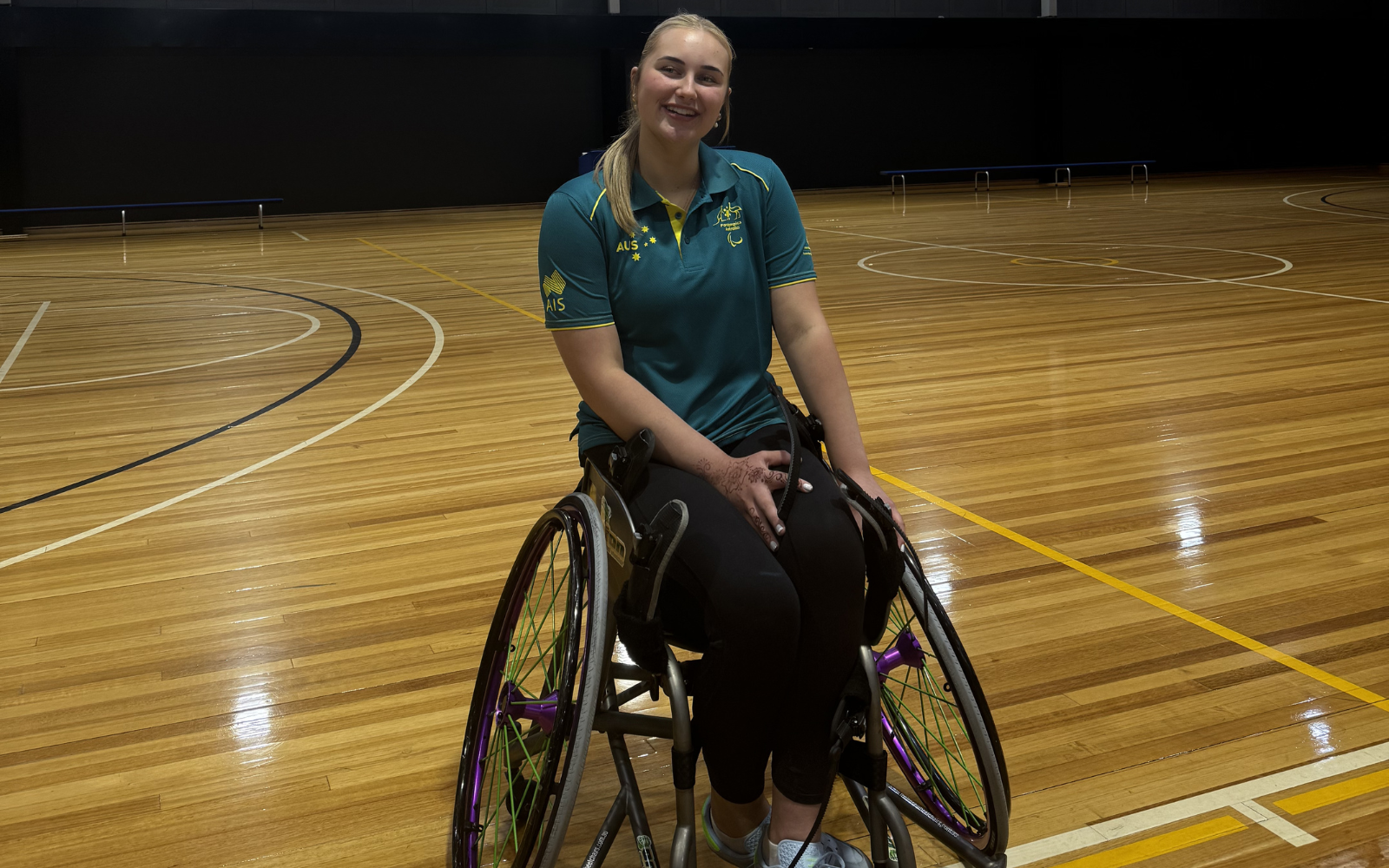 Smiling woman in a wheelchair on a polished gym floor, wearing a teal polo shirt and black leggings.