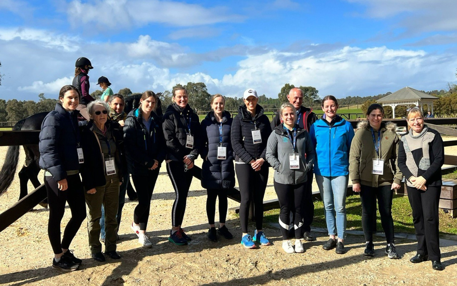 Group of people posing for a photo at an outdoor equestrian facility, wearing jackets and name badges, with a pony behind them on a fence.