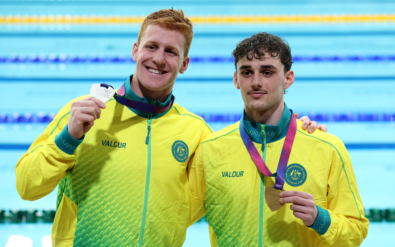 Two young swimmers in bright yellow jackets pose and smile, each wearing a medal around the neck near a pool backdrop.