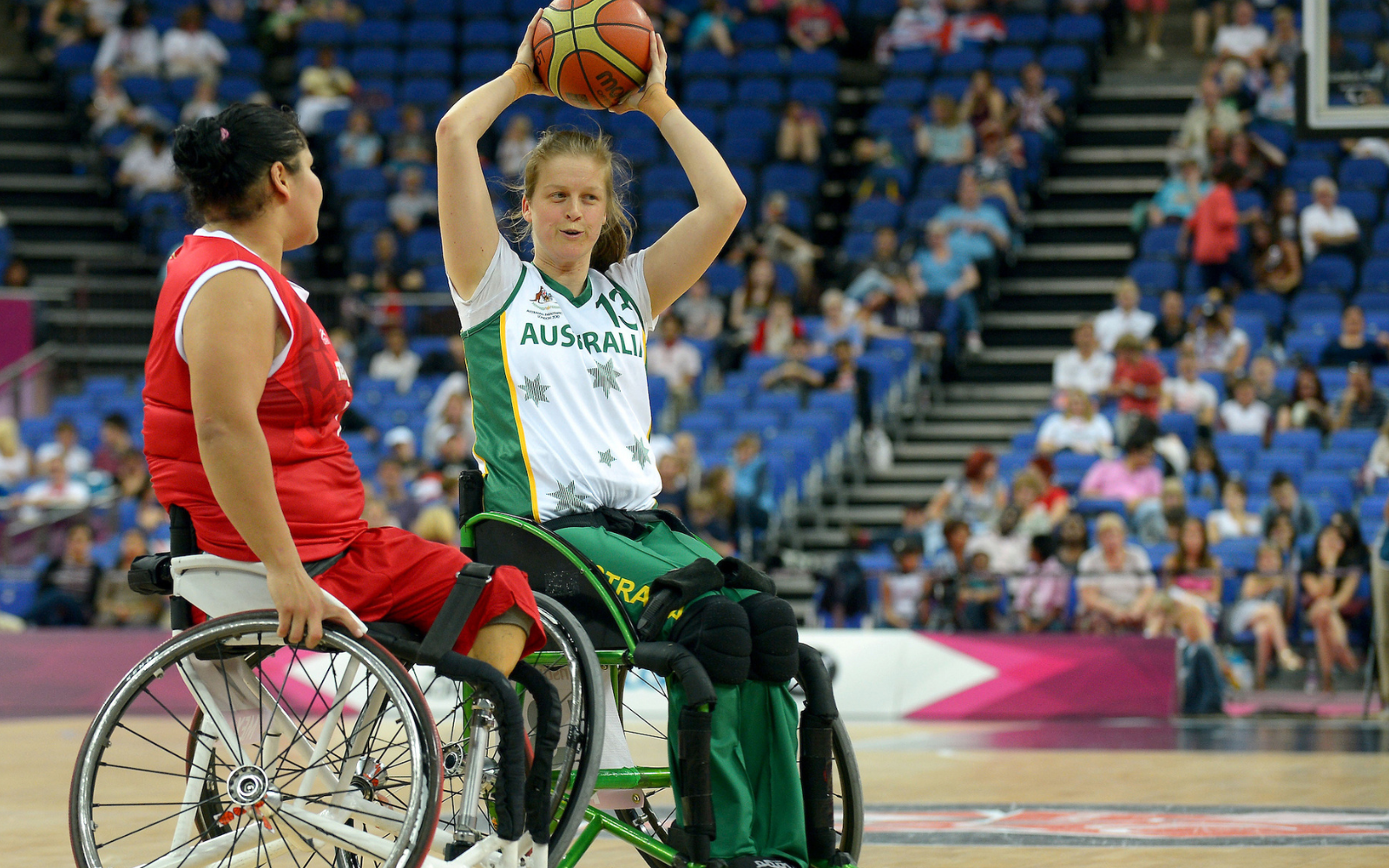 Female wheelchair basketball players in action during a game; one in white Australia jersey raises the ball while the opponent in red watches, crowd in bleachers behind them.