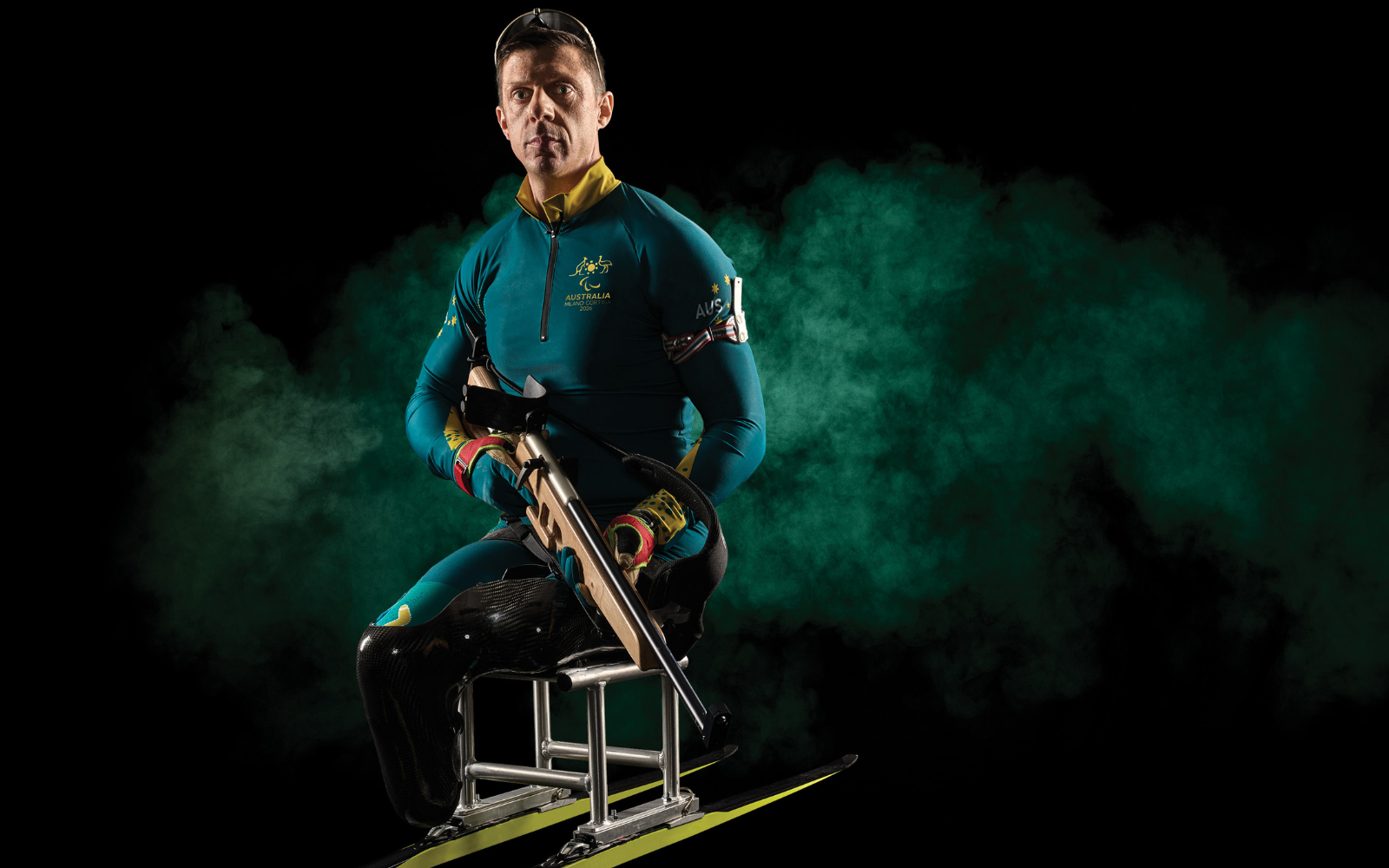 Male biathlete in teal Australia uniform seated on a skis setup, holding a rifle against a black background with green smoke.