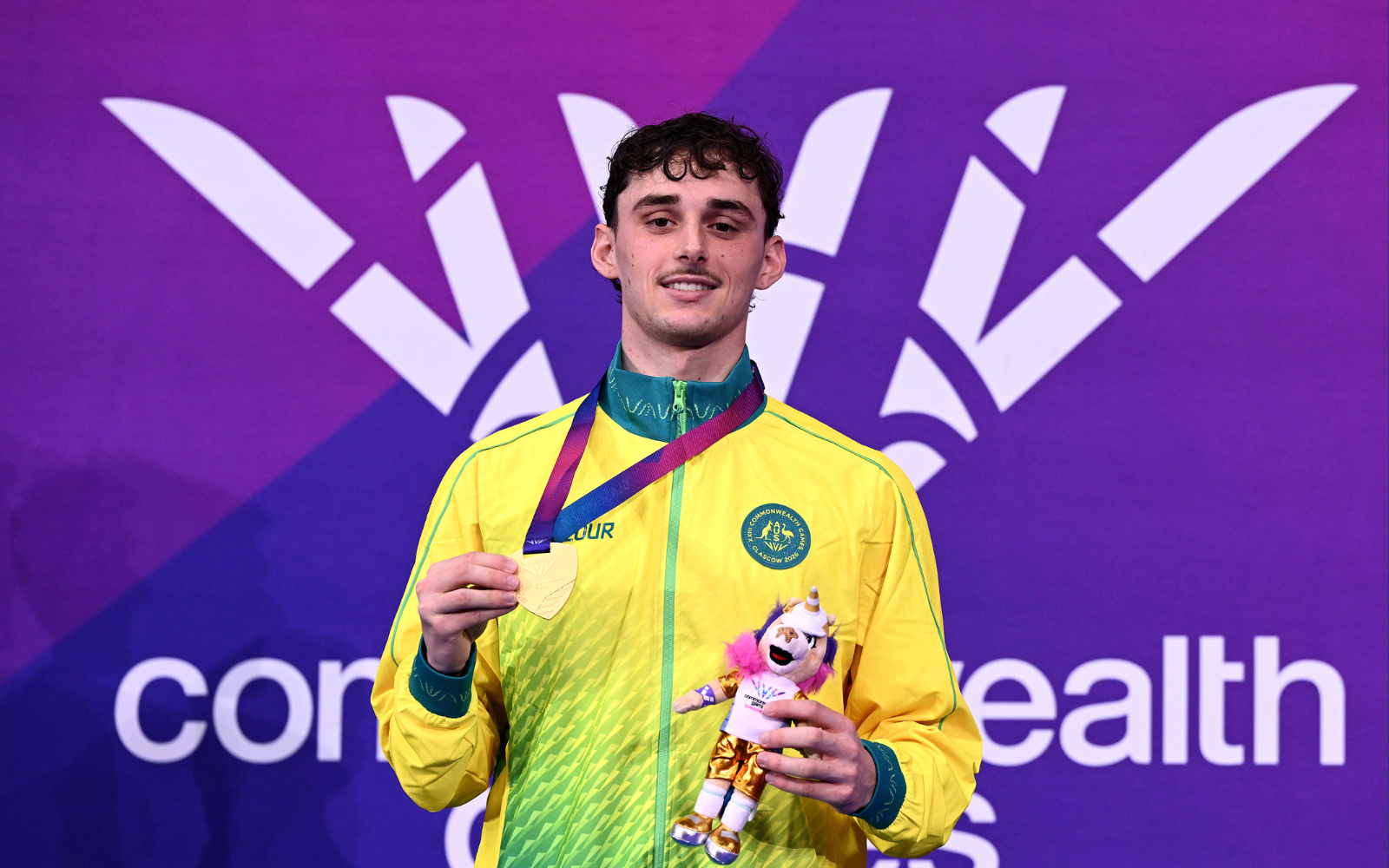 Young athlete on a podium holding a gold medal and a unicorn plush, wearing a yellow‑green jacket against a purple backdrop with event branding.