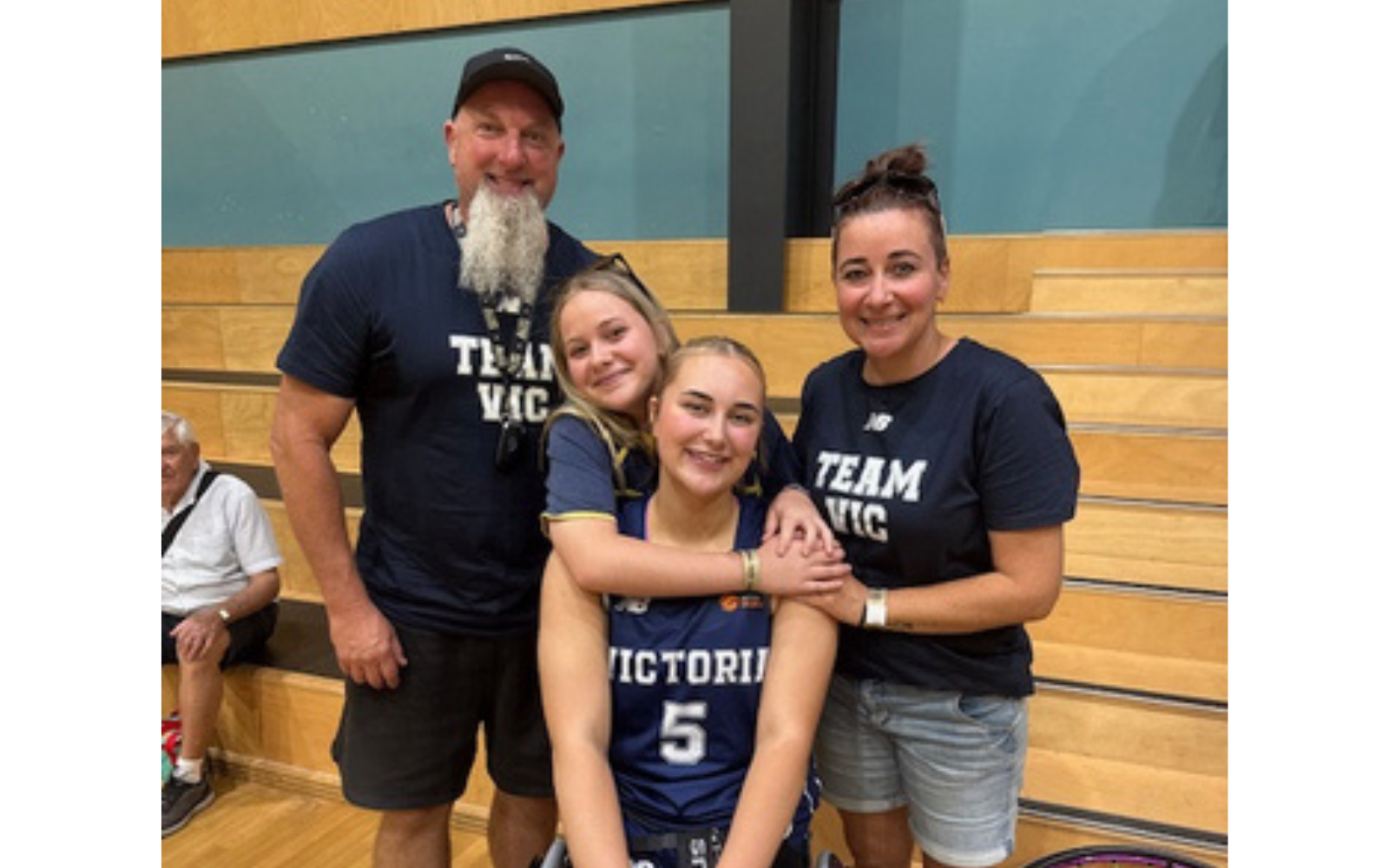 Group photo of four teammates in a gym: a bearded man in a navy 'TEAM VIC' shirt stands left, two women stand beside him, one seated in a wheelchair in front wearing a blue jersey.