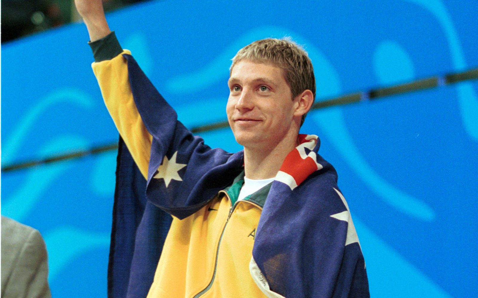 Athlete with a yellow-blue jacket draped in an Australian flag raises his hand in victory against a blue background.