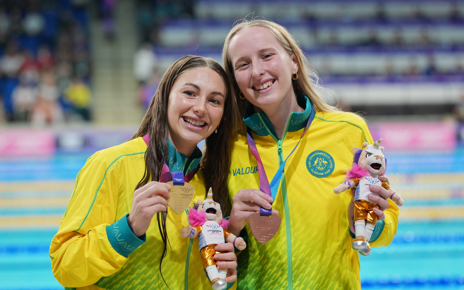 Two smiling female swimmers in yellow jackets pose with medals and plush mascots at a swimming event.