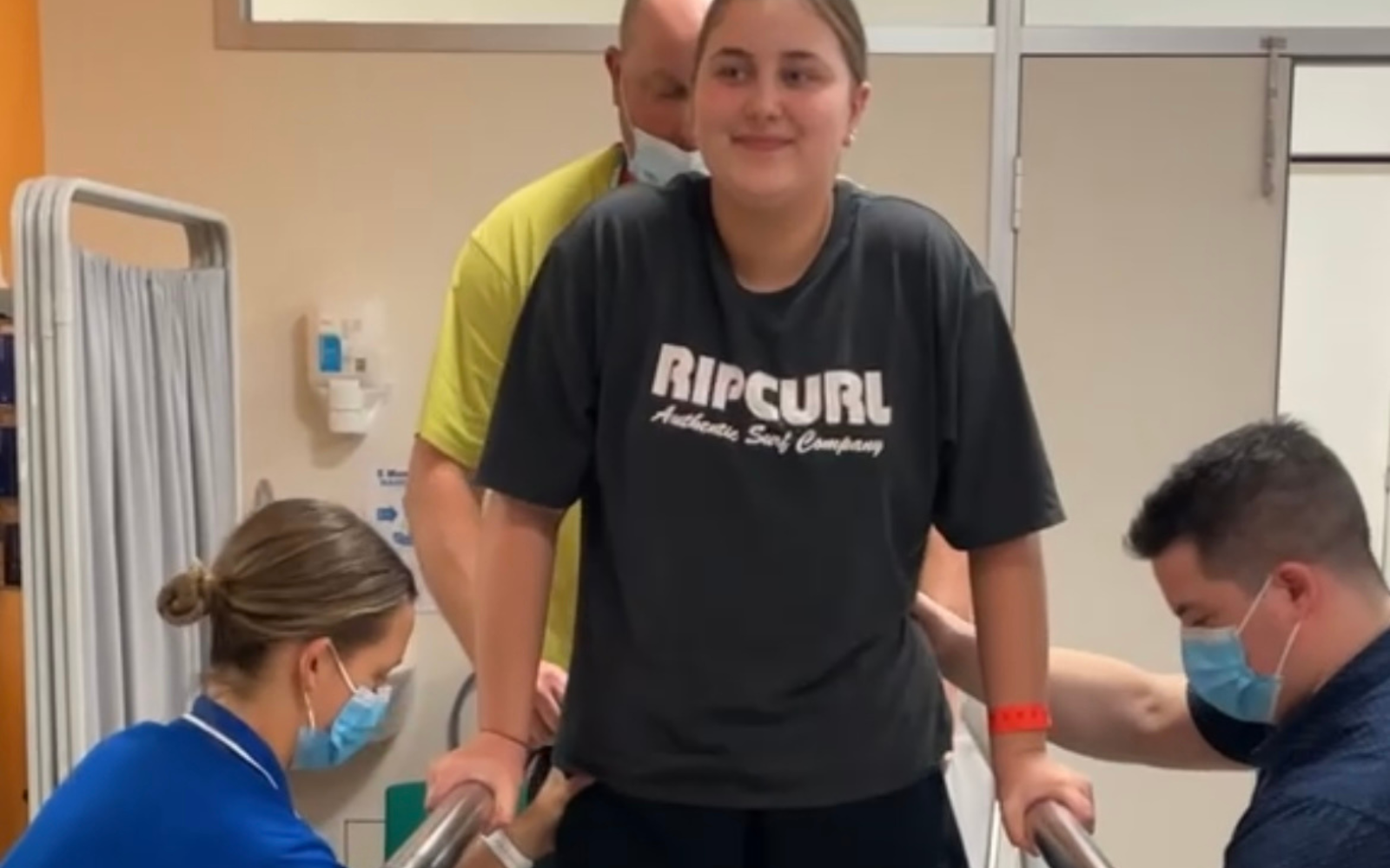 Young patient in a dark RIP CURL shirt stands on parallel bars and is supported by two masked therapists in a clinical rehab room.