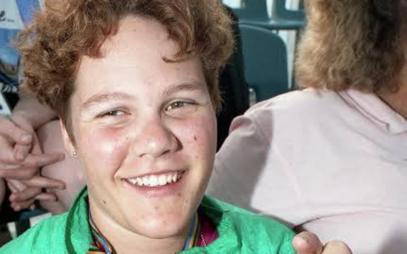 Smiling person with short red hair wearing a green jacket, looking at the camera outside in daylight.