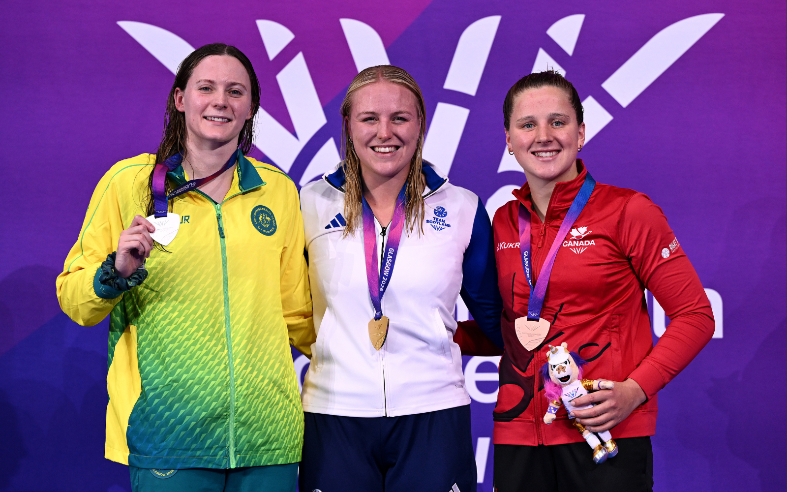 Three female athletes stand on a podium, each wearing a medal: silver on the left, gold in the center, bronze on the right, with a purple backdrop and event branding behind them.
