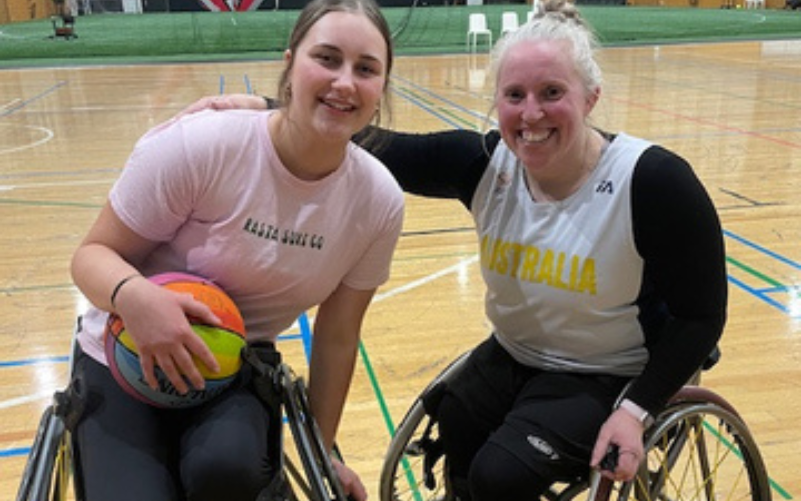 Two women in wheelchairs posing on a gym court, one holding a colorful ball and smiling at the camera.