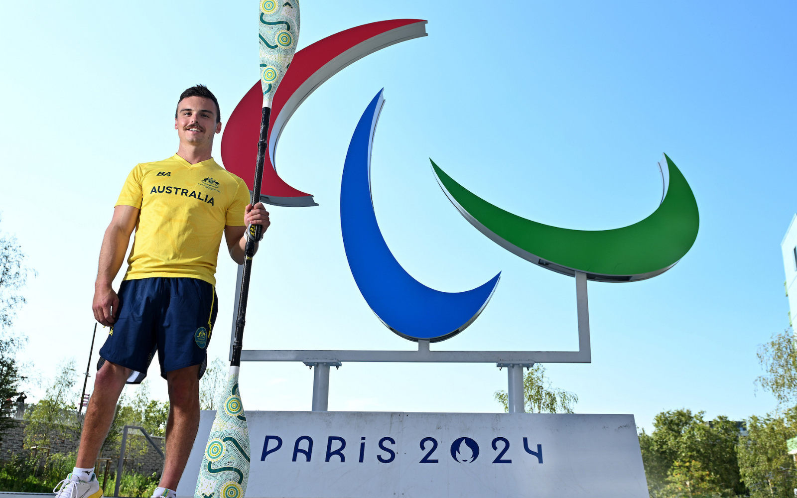 Athlete in a yellow Australia shirt stands with a decorative paddle in front of the Paris 2024 sculpture and sign.