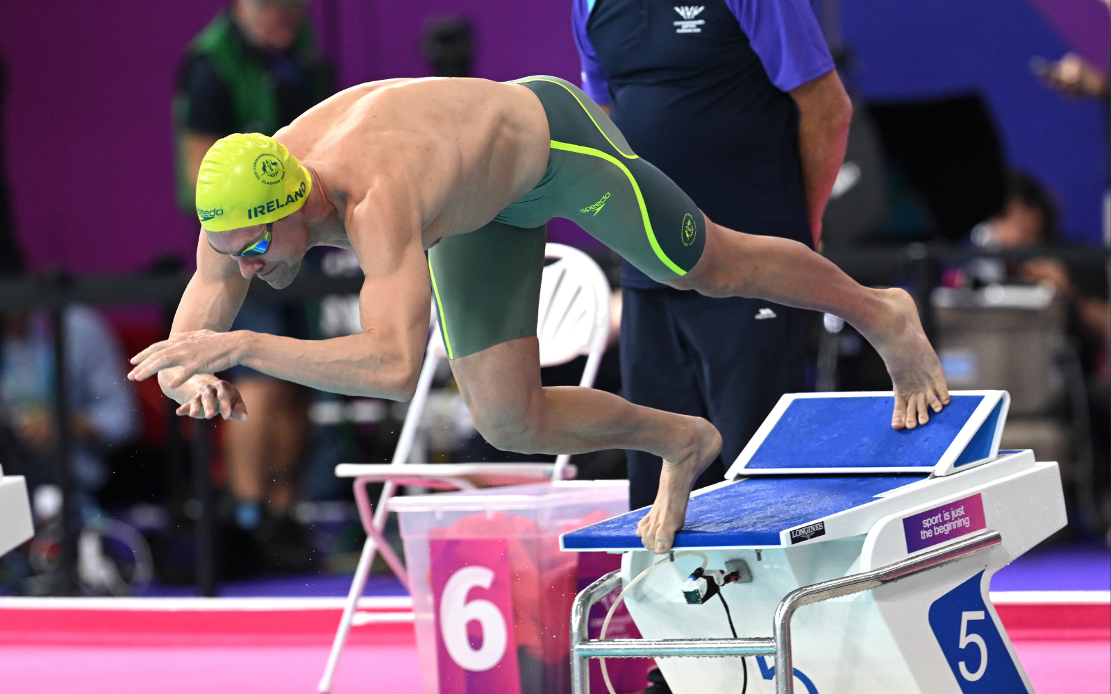 Swimmer in a bright yellow cap diving off the starting block during a race in a pink pool area, Ireland on cap, green suit with neon trim