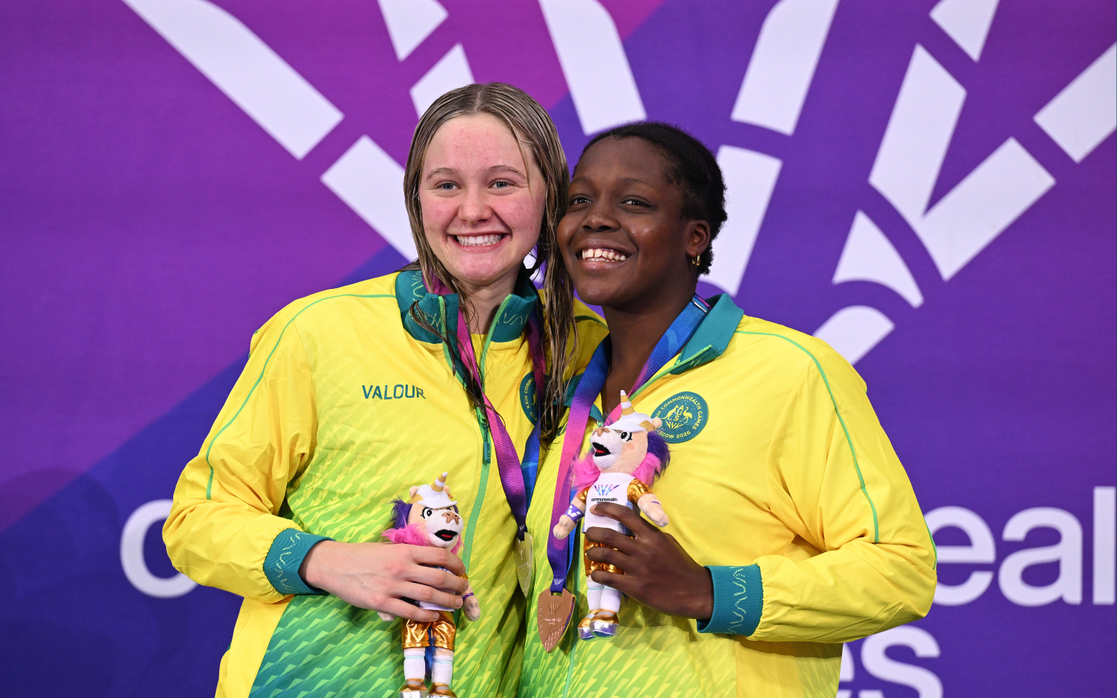 Two female athletes in yellow-green jackets smile with medals and unicorn plush toys on a purple backdrop, celebrating a podium finish.