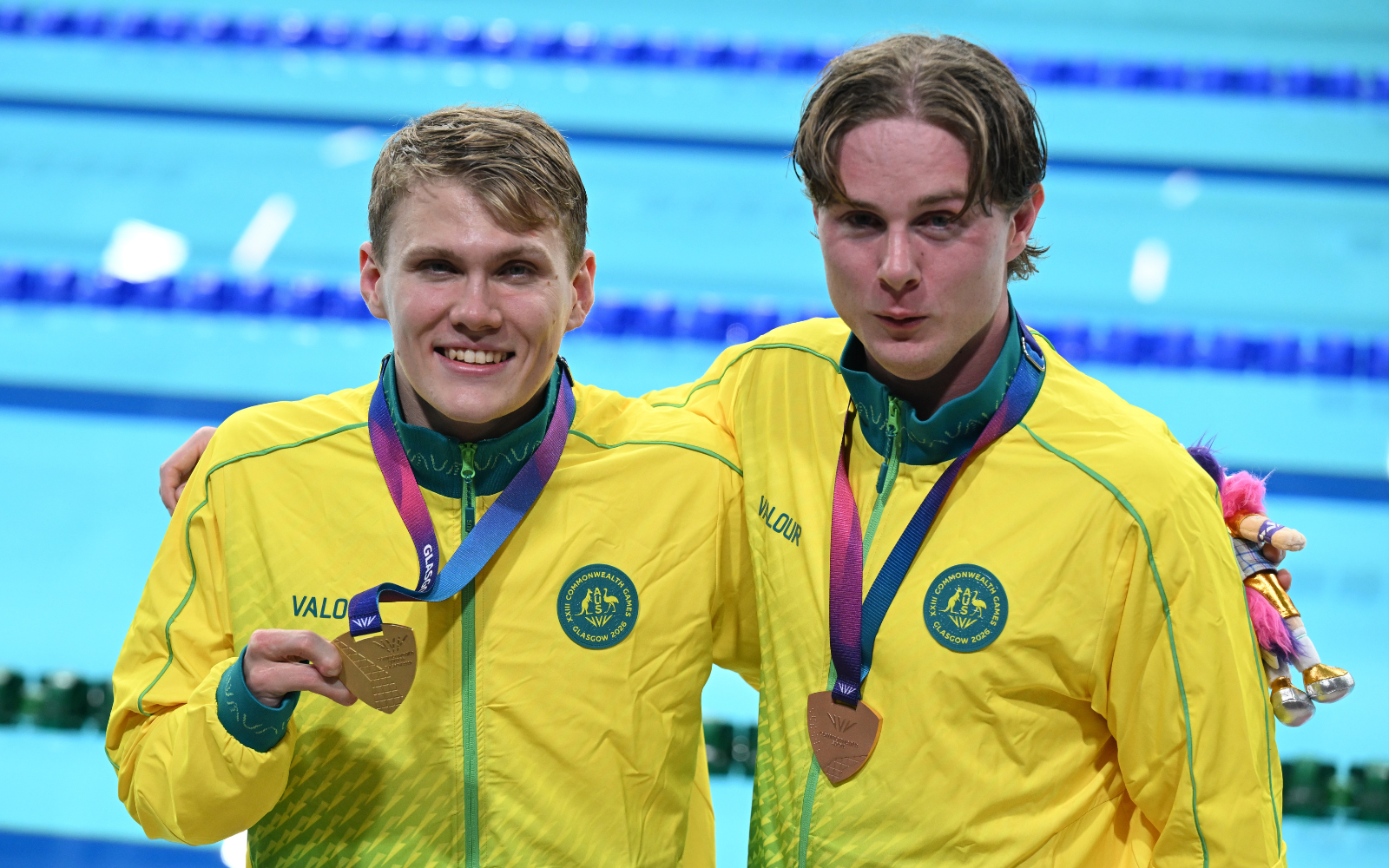 Two male swimmers in yellow jackets, smiling at the pool, with bronze medals on purple ribbons around their necks.
