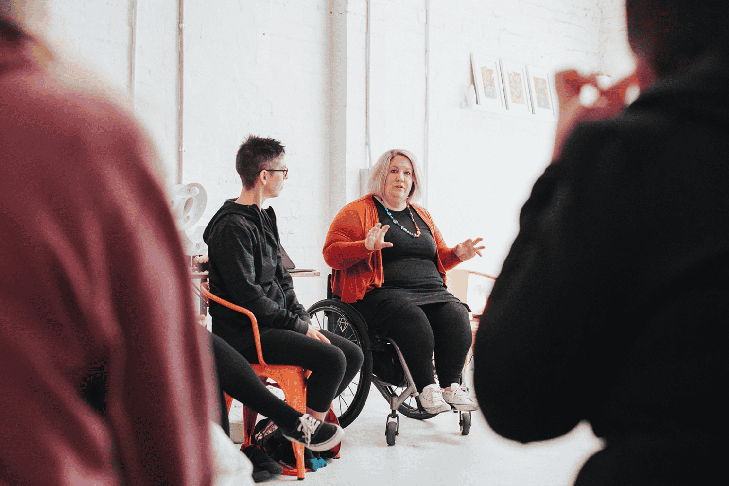 Woman in a wheelchair gesturing while speaking to a small group in a bright, minimalist room; a young man in an orange chair listens beside her.