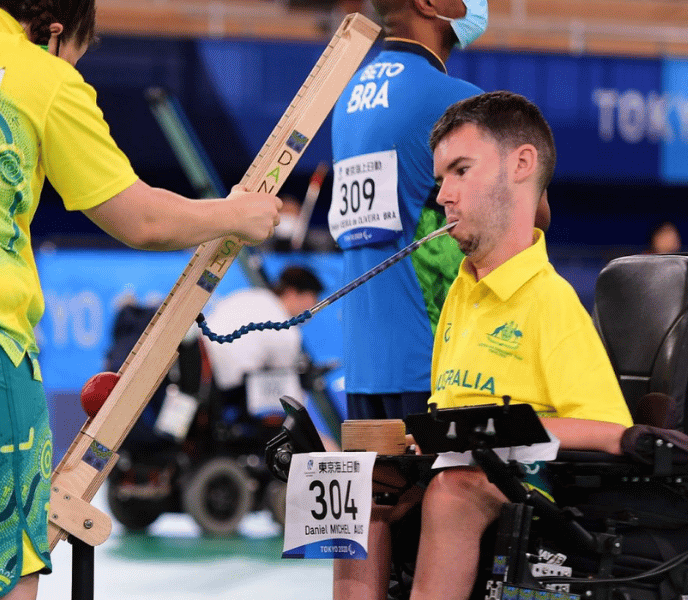 Paralympic athlete in a yellow Australia jersey sits in a wheelchair during a track-and-field event, with a teammate in blue nearby and a wooden measurement device in view.