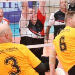 Volleyball match in progress; two yellow-jersey players (#3 and #6) on the floor near the net with black uniforms nearby.