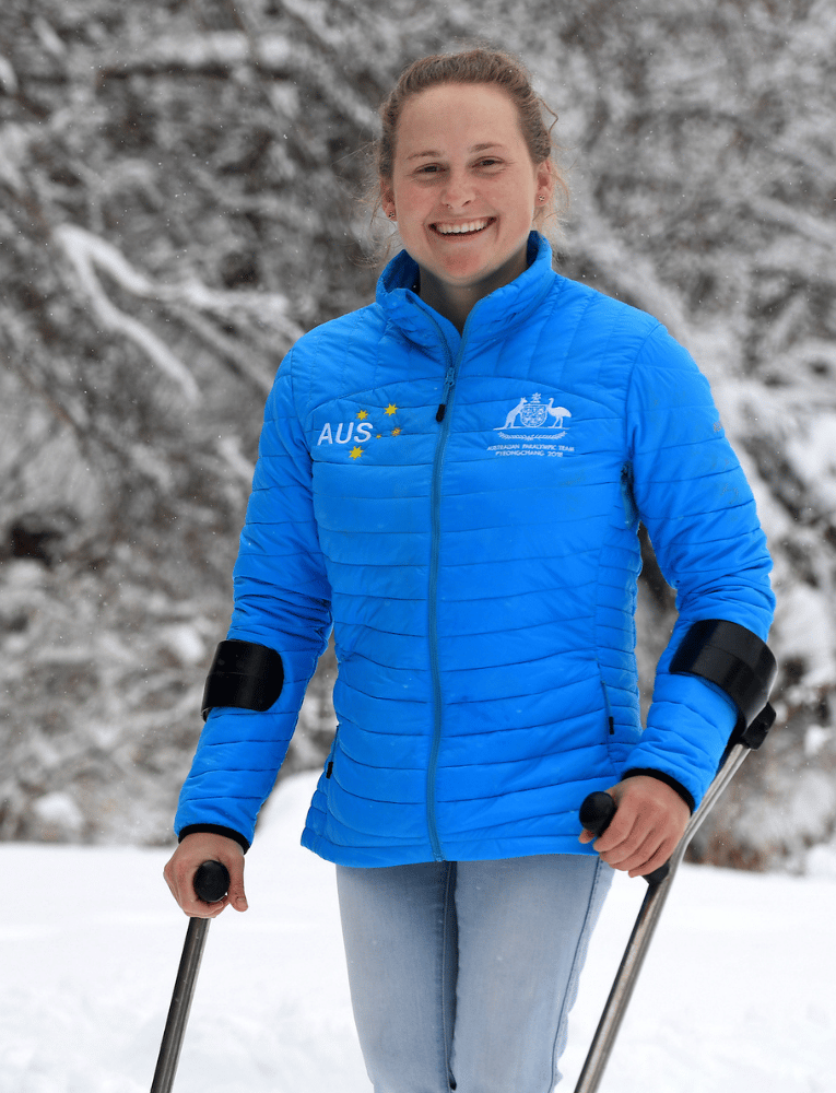 Smiling woman in a bright blue quilted jacket using ski poles in a snowy forest setting.