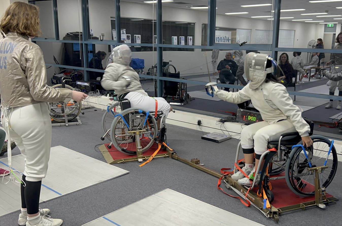Two wheelchair fencers in white protective gear duel on a fencing piste, with timing equipment nearby and spectators watching through a glass barrier.