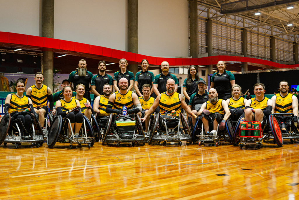 Group portrait of a wheelchair basketball team in yellow and black uniforms posing on an indoor court, with backpacks and equipment nearby.