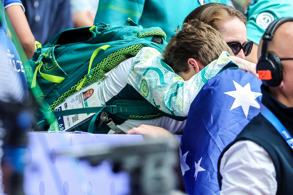 Athlete with a teal backpack bending over a barrier, wearing an event ID badge on a lanyard.