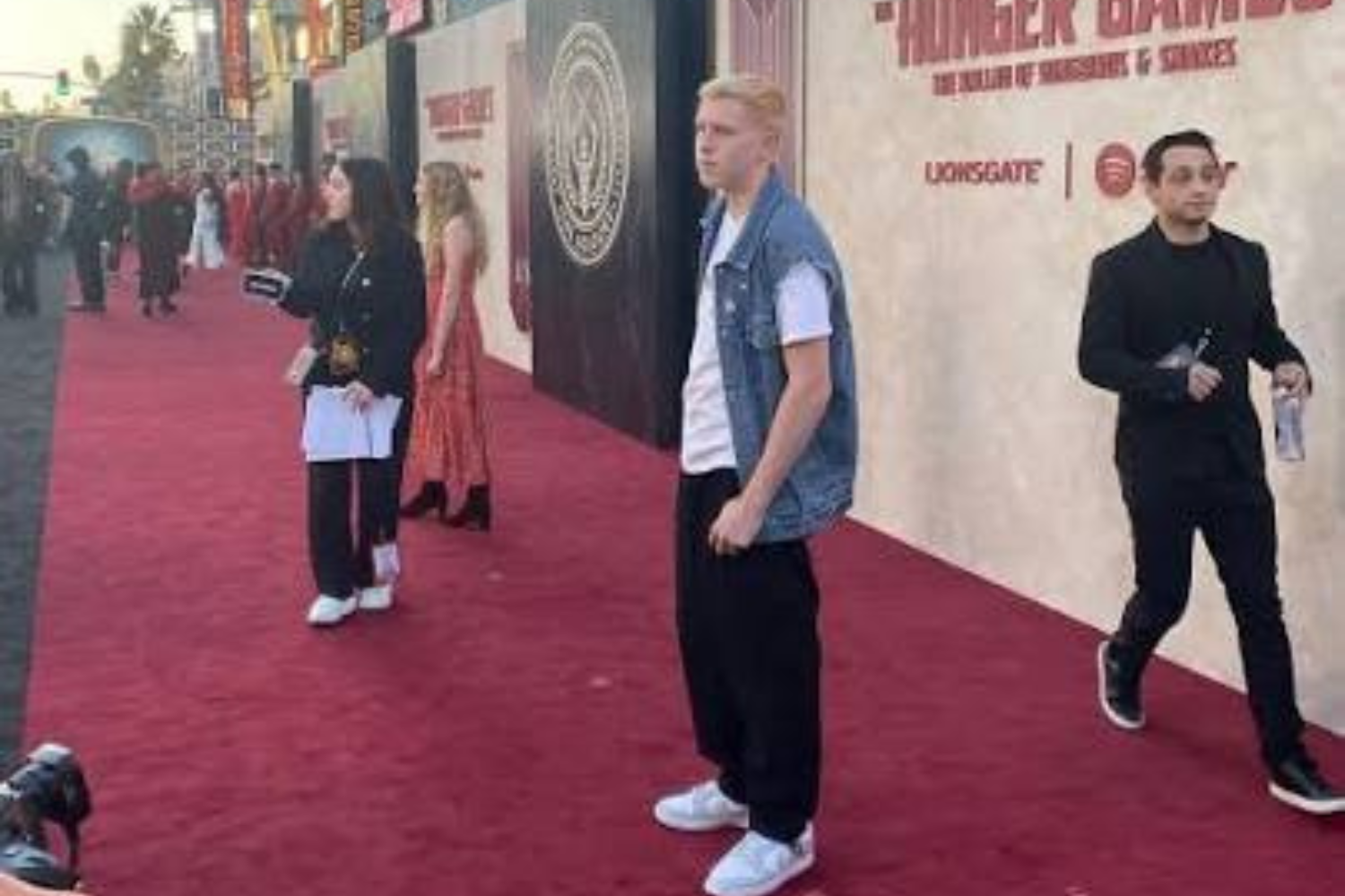 Celebrities pose on a red carpet against a backdrop, with a man in a denim vest and white sneakers in the foreground.