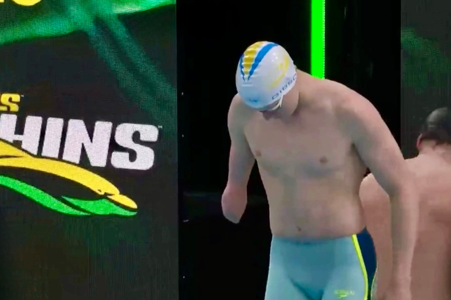 Swimmer in blue-and-neon trunks stands at the pool edge, wearing a white cap with blue and yellow stripes, preparing to dive.