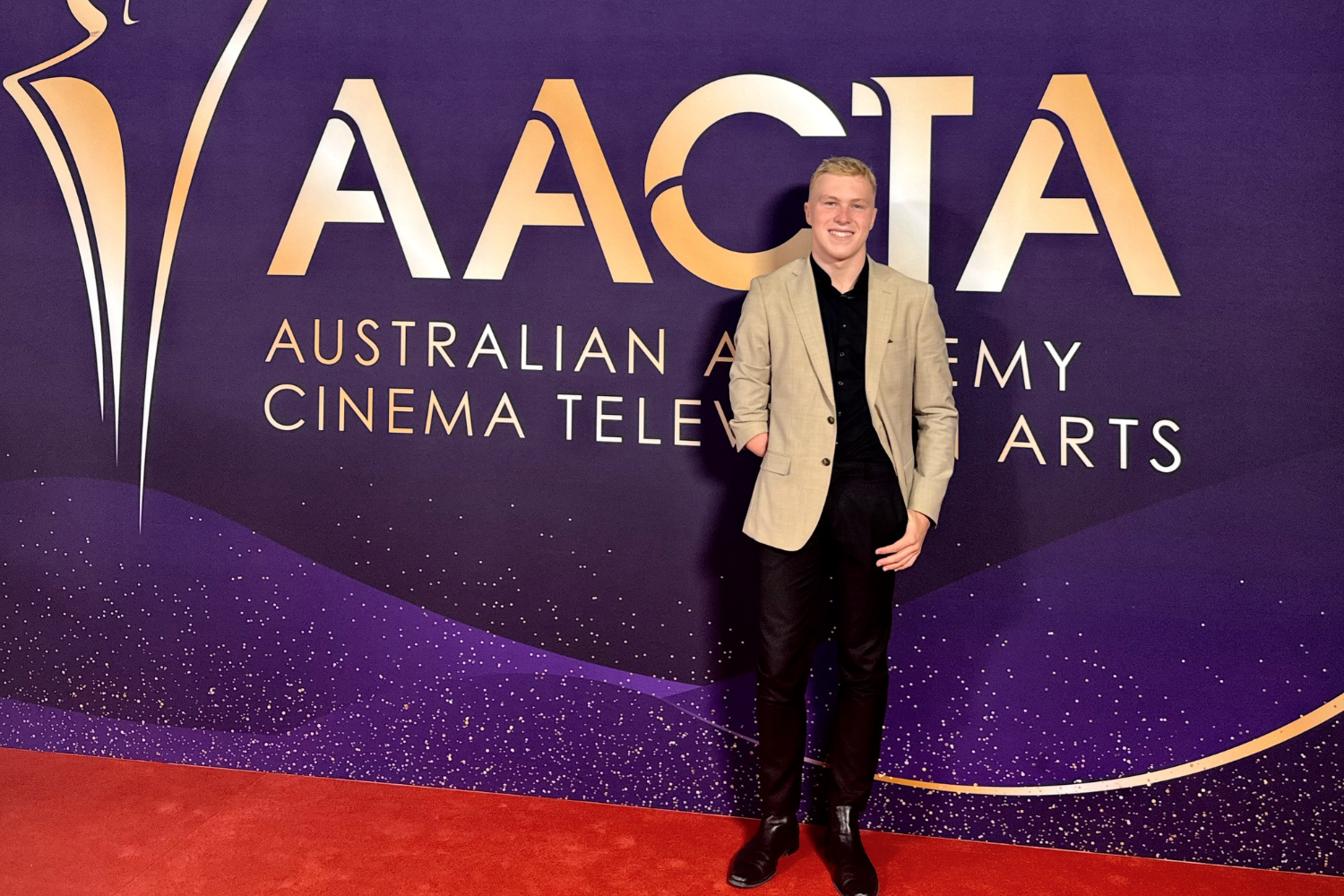 Man posing on a red carpet in front of a purple AACTA backdrop with gold lettering.