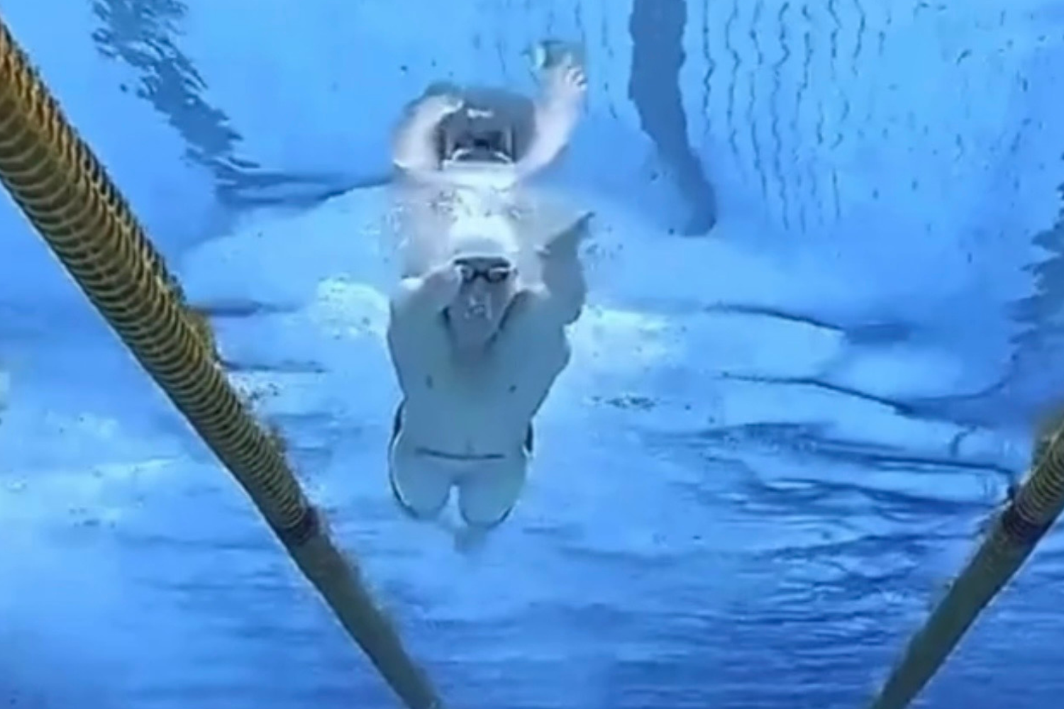 Underwater swimmer in a pool faces the camera with one arm raised and the middle finger extended toward the lens, lanes visible in the background.