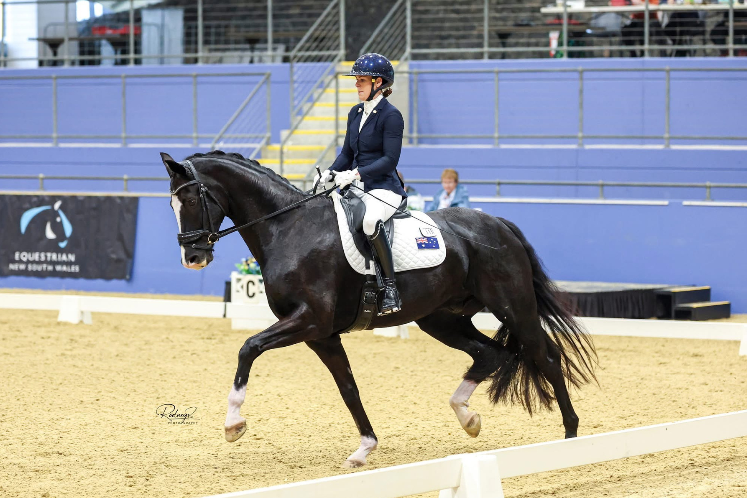 Female equestrian in navy jacket and helmet riding a black horse in a dressage arena, performing a collected trot with white saddle pad and gloves.