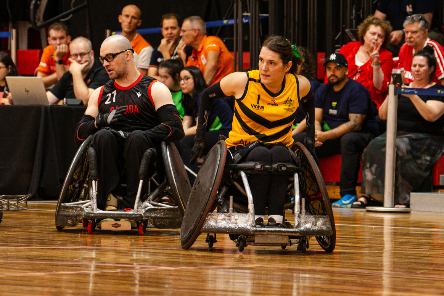 Two wheelchair basketball players in action on a wooden court, one in black Canada jersey and the other in yellow and black, with spectators behind them