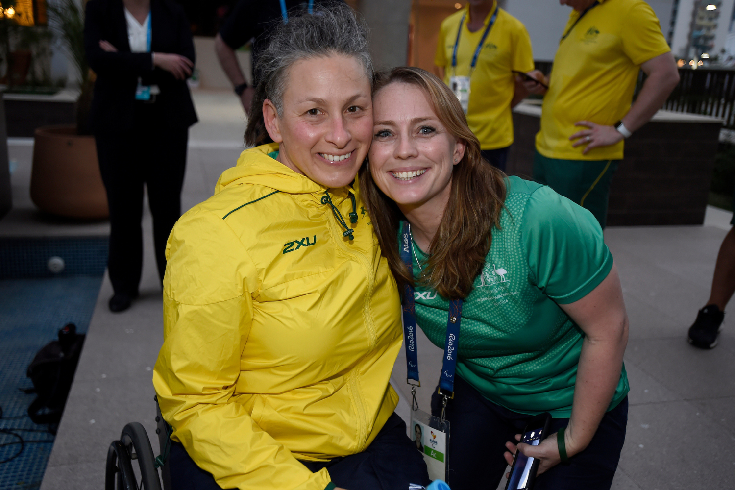 Two women smiling at the camera, one in a yellow jacket and the other in a green shirt, posing together at an outdoor event with lanyards visible around their necks.