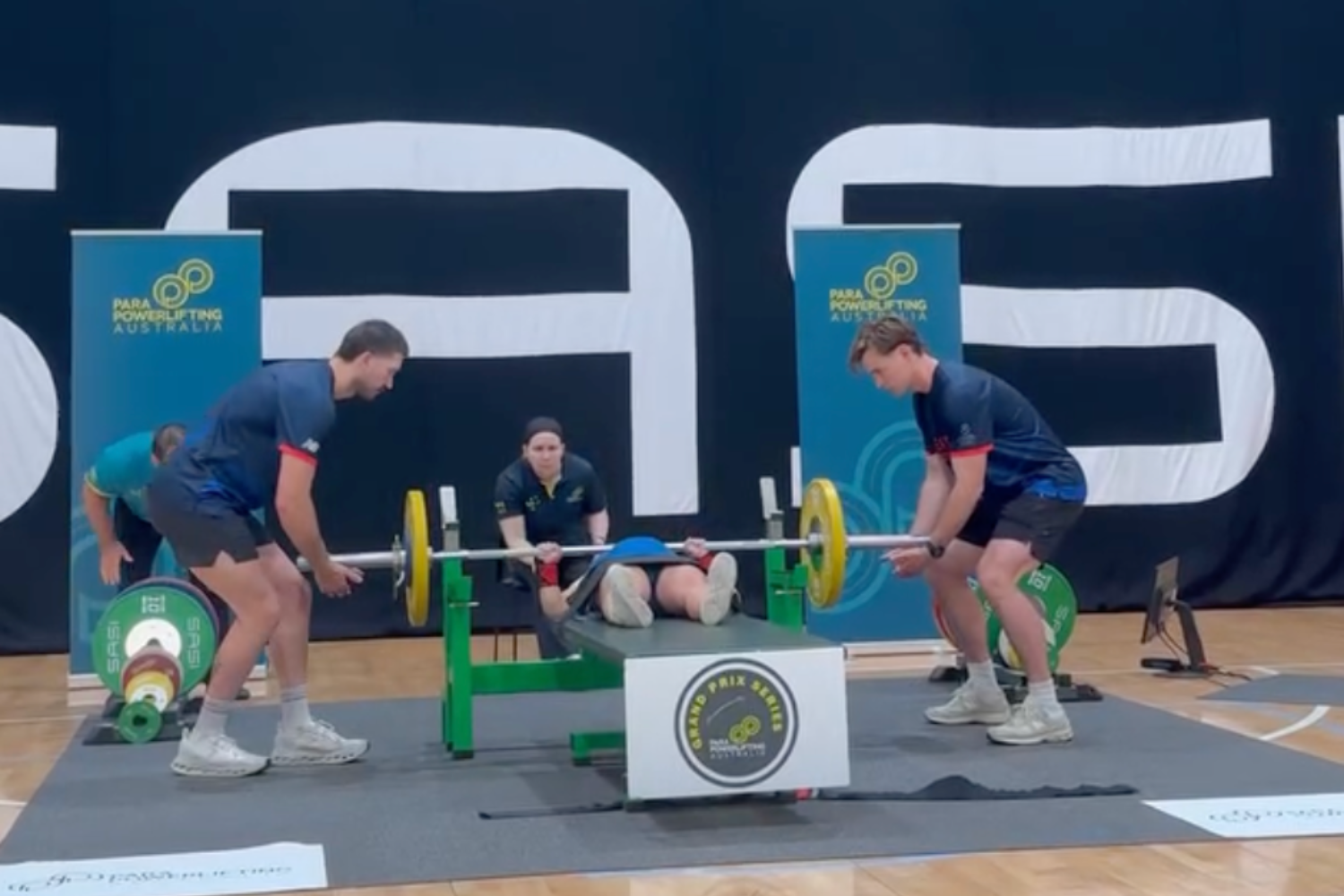 Athletes perform a bench press at a para powerlifting competition, with spotters on each side and a referee seated behind the bench by banners that read ‘Para Powerlifting Australia’.