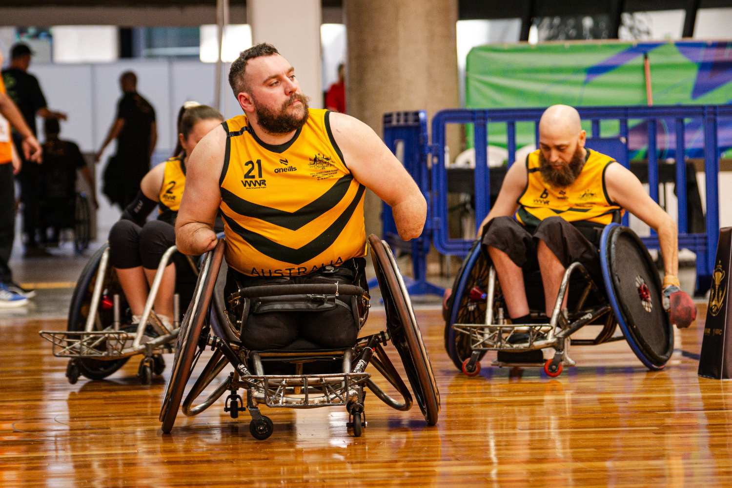 Wheelchair rugby players in yellow jerseys race across a polished gym floor, with a bearded player in the foreground pushing his chair forward in action.