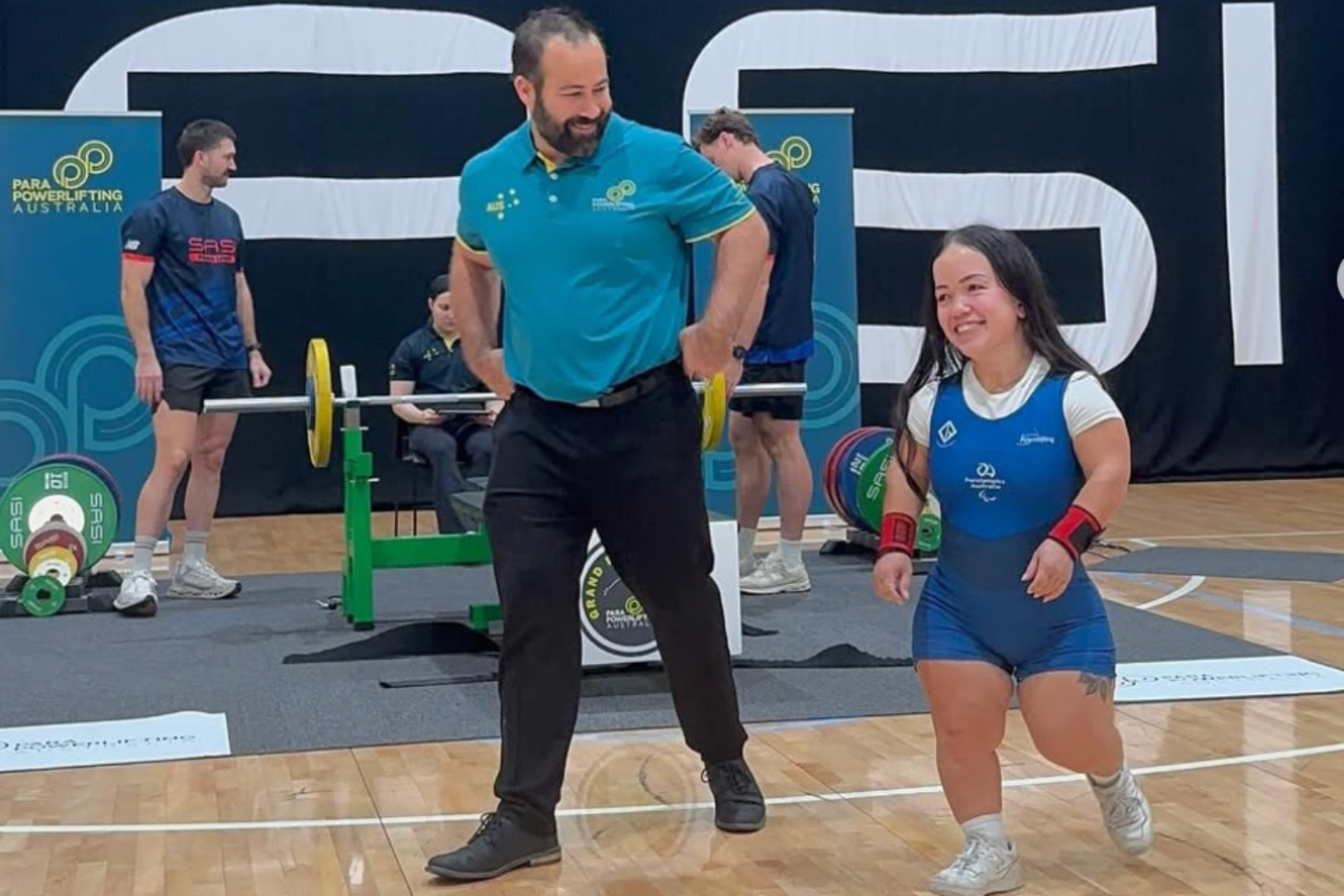 Powerlifting competition scene: man in teal shirt stands with hands on hips on the platform as a smiling female lifter in a blue singlet runs toward the camera in the foreground.