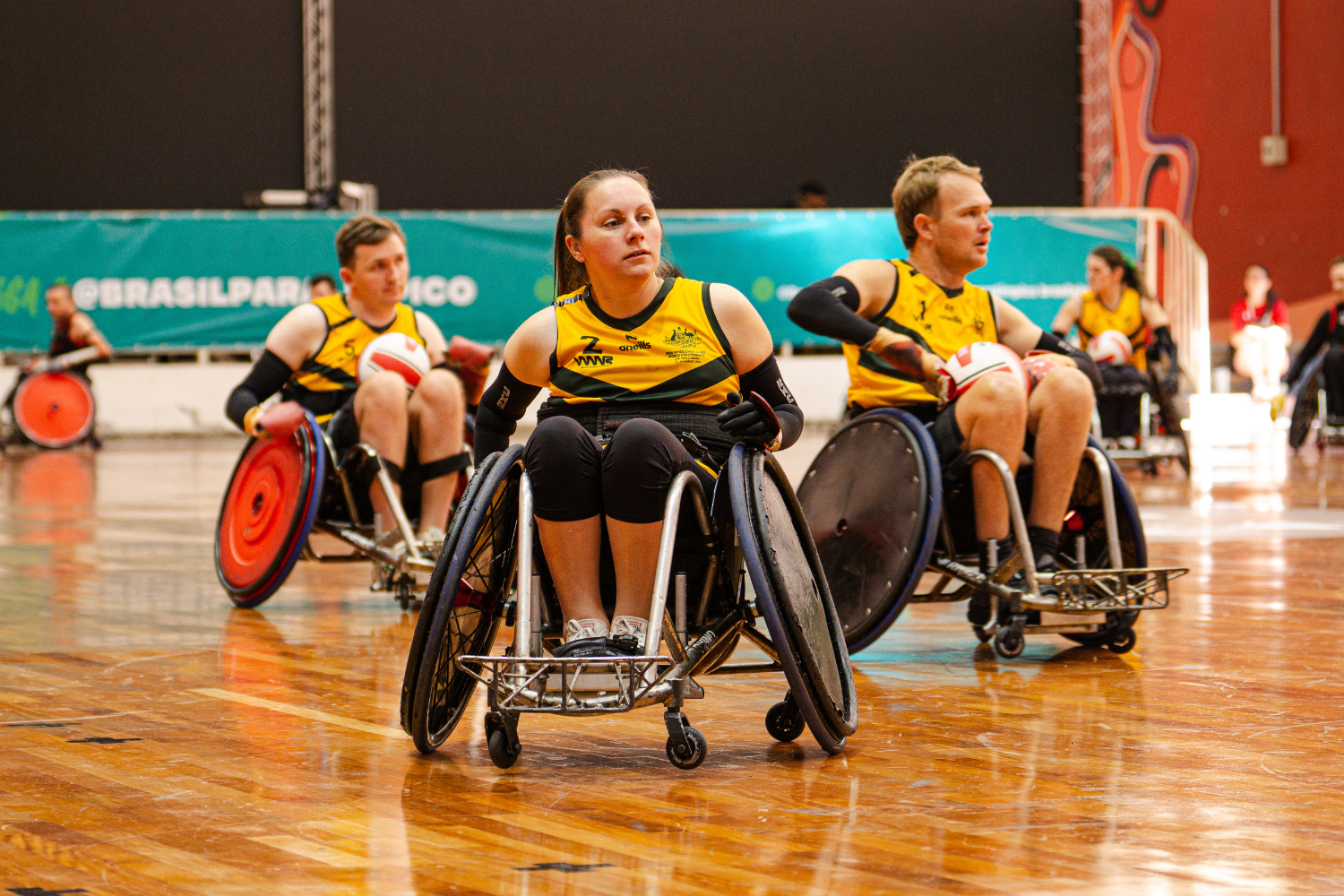 Wheelchair rugby players in yellow jerseys compete on a polished indoor court; a central female player in a wheelchair looks ahead as the action unfolds in the foreground.