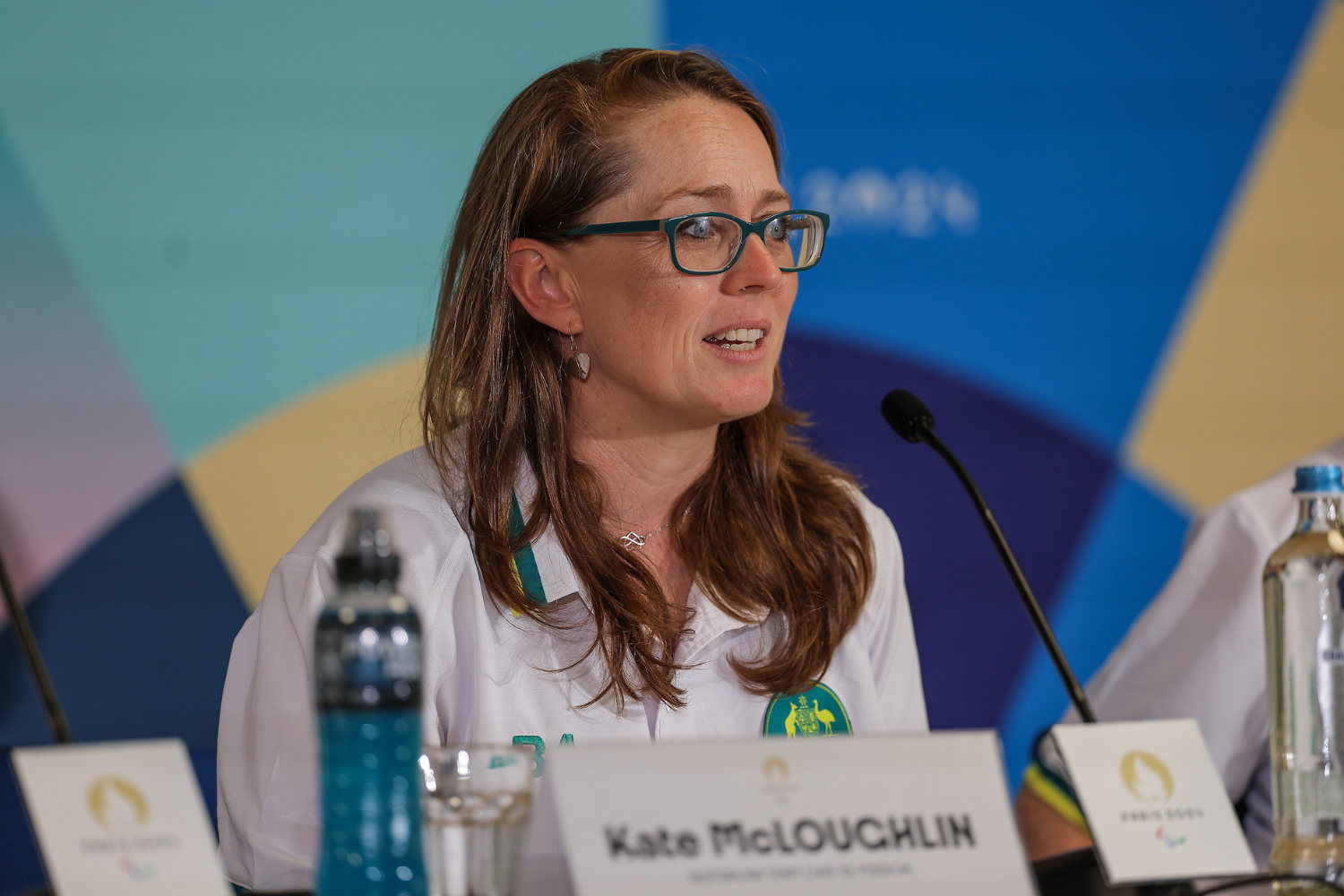 Panelist Kate McLoughlin speaks into a microphone at a conference, with a colorful geometric backdrop and water bottle on the table on her left