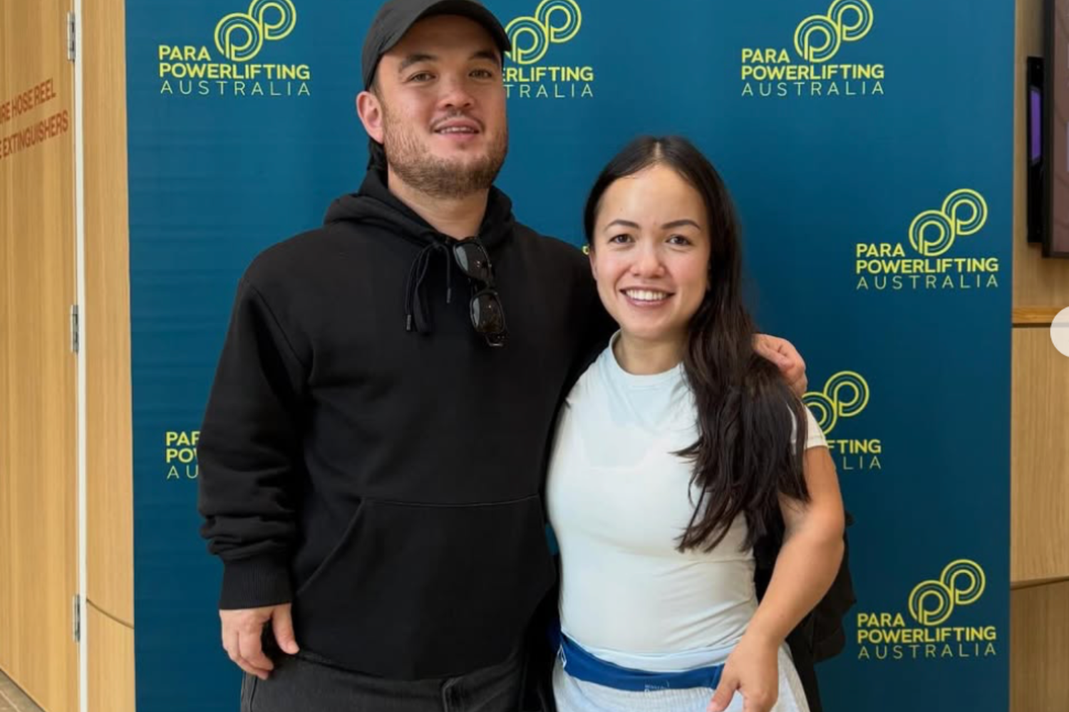 Two people pose for a photo in front of a blue Para Powerlifting Australia backdrop; a man in a black hoodie and cap stands with a smiling woman in a light blue top.