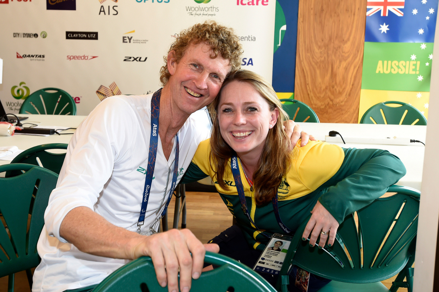 Two smiling adults posing together at an event, wearing lanyards, with sponsor logos on a backdrop behind them.