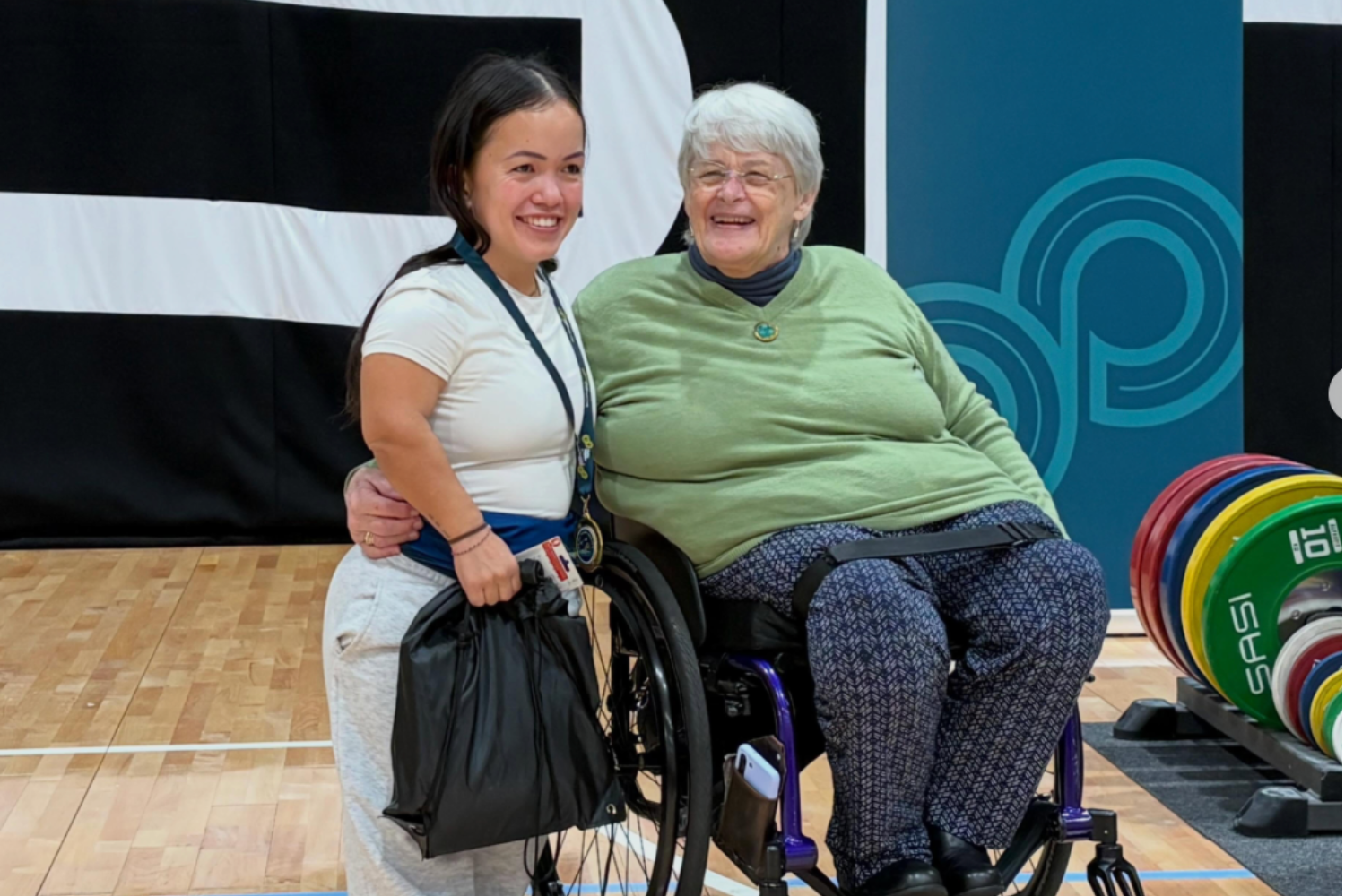 Smiling young woman helping an older woman in a wheelchair at a gym, with weight plates visible in the background.